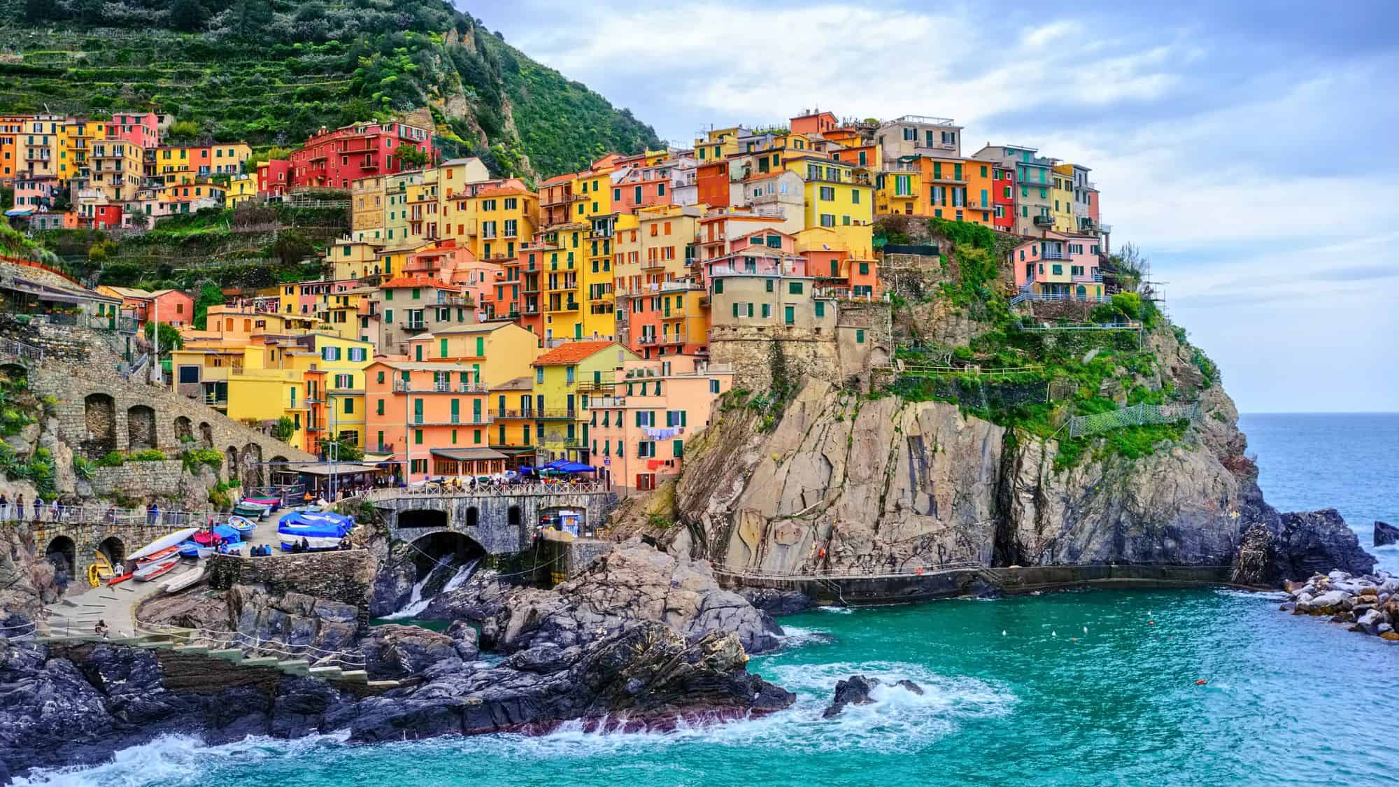 A vibrant Cinque Terre village with colorful houses clinging to cliffs above the turquoise sea, surrounded by terraced vineyards.