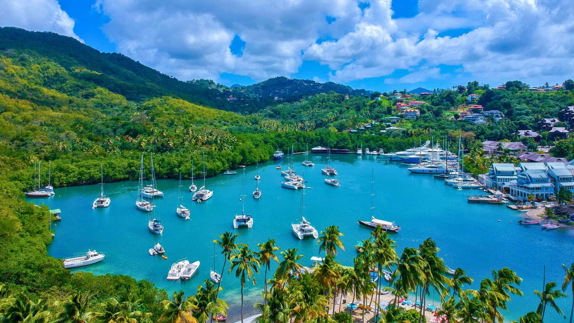 Aerial view of Marigot Bay in St. Lucia, with turquoise waters dotted by sailboats and catamarans, framed by vibrant green hills and coastal villas.