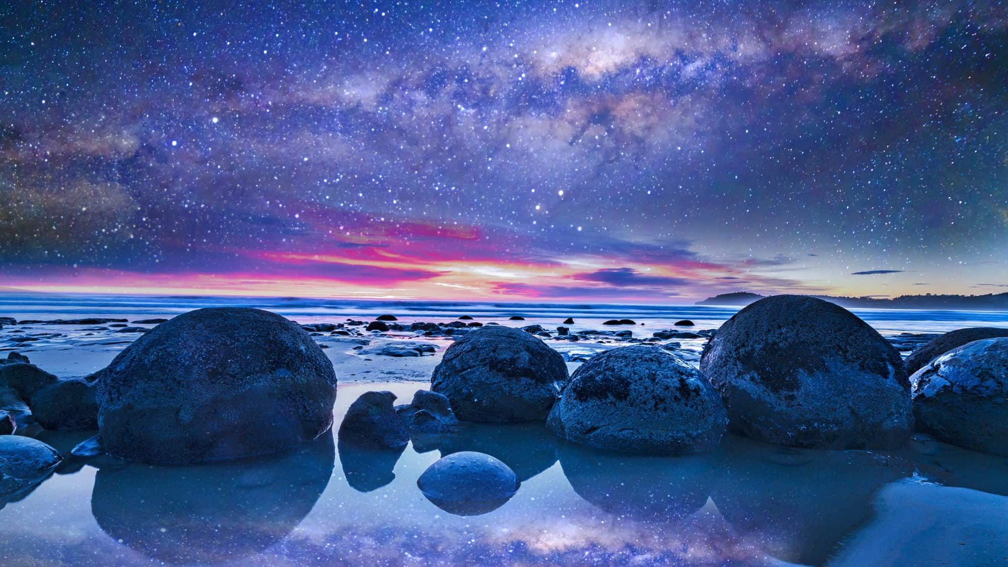Large, spherical boulders rest along the shore under a vivid, star-filled night sky with the Milky Way visible above and hues of purple, blue, and pink at the horizon. The reflective tide pools mirror the sky’s colors, adding to the surreal, dreamlike feel.