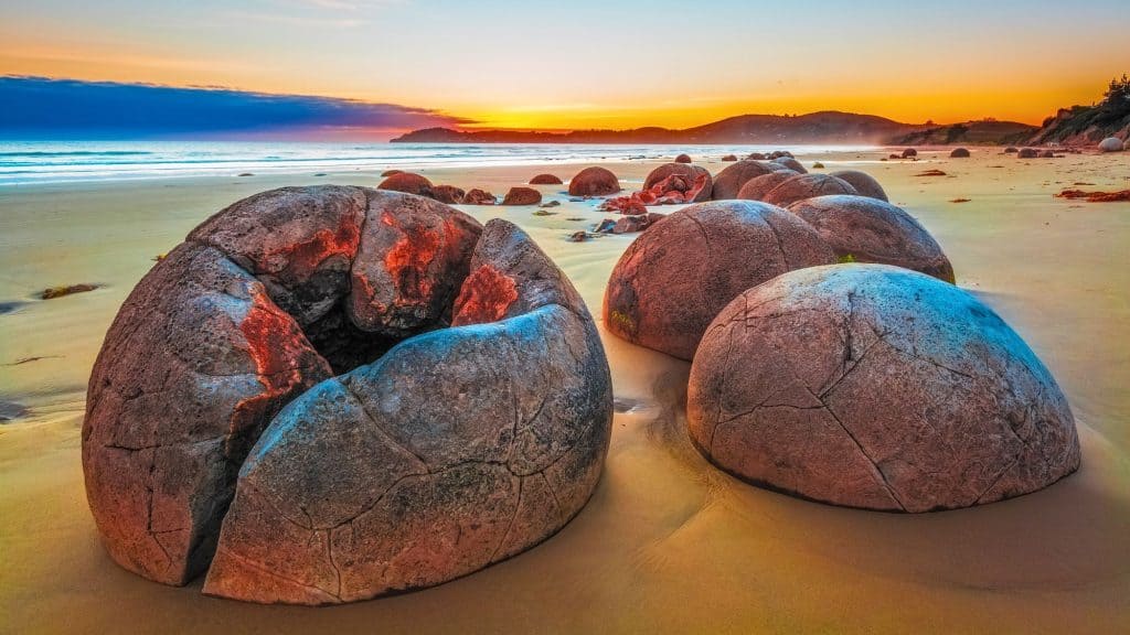 Smooth, spherical Moeraki Boulders lie scattered along a golden beach in New Zealand, glowing with hues of red and blue as the first light of sunrise illuminates the scene. The cracked surfaces and unique formations add to the mystique of this coastal geological marvel.