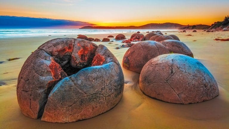 Smooth, spherical Moeraki Boulders lie scattered along a golden beach in New Zealand, glowing with hues of red and blue as the first light of sunrise illuminates the scene. The cracked surfaces and unique formations add to the mystique of this coastal geological marvel.