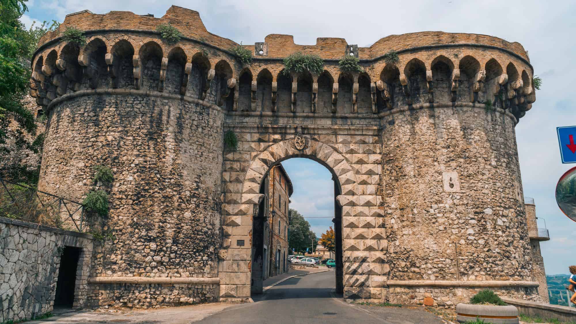 A massive, medieval stone gate flanked by round towers in Narni, Italy, featuring a pointed arch and battlemented top with tufts of greenery.
