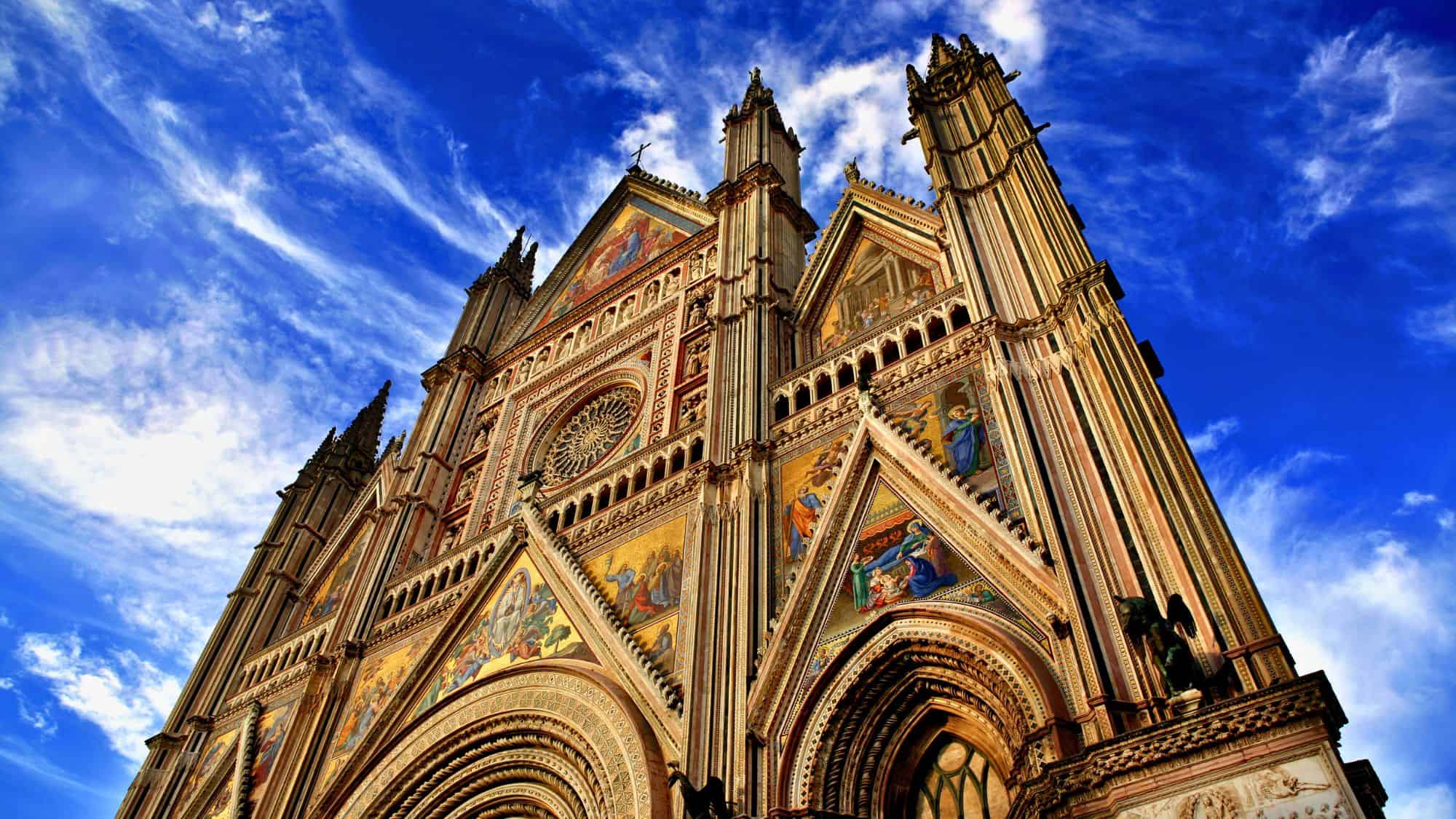 Upward view of the ornate facade of Orvieto Cathedral, rich with colorful mosaics, Gothic spires, and intricate detailing set against a vivid sky.