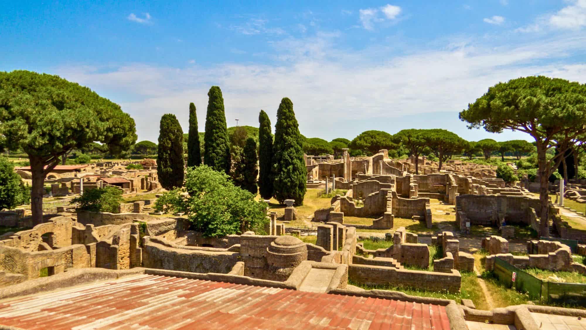Ancient Roman ruins surrounded by tall cypress and pine trees, with crumbled stone walls and paths weaving through the archaeological site.