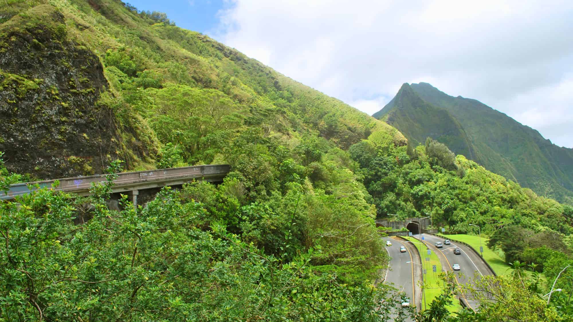 Lush, green mountain landscape surrounds the Pali Highway tunnels on Oʻahu, with traffic winding through dense tropical foliage.