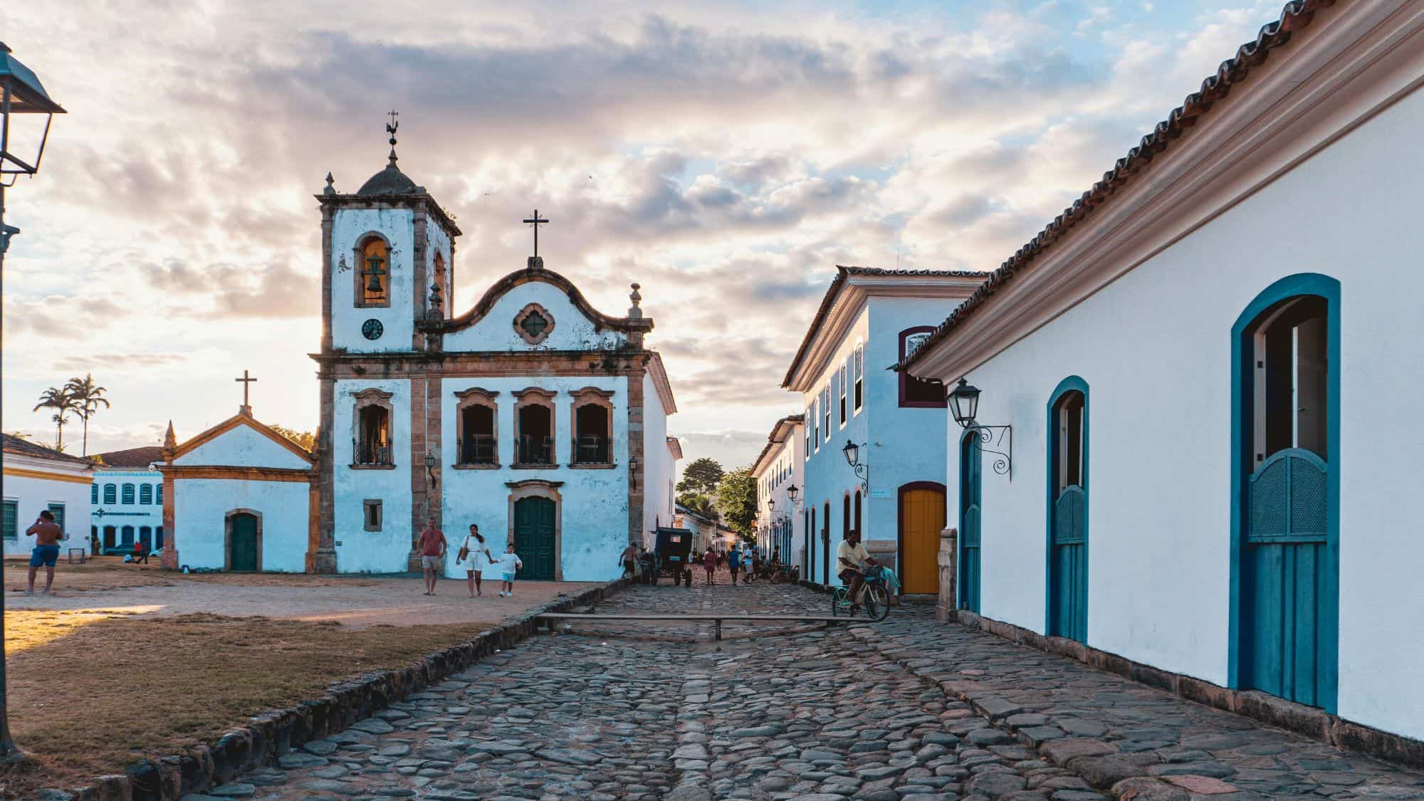 Cobblestone streets lead to a whitewashed colonial church flanked by historic homes with bright blue accents in Paraty, Brazil. The golden-hour lighting and classic architecture give the scene a timeless, picturesque charm.