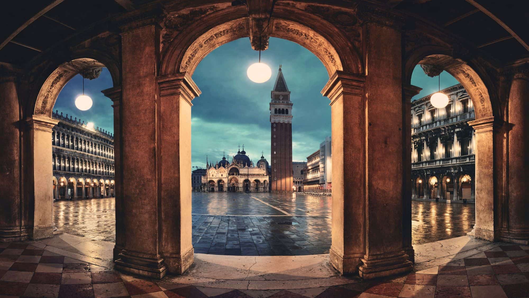 A dreamy nighttime view of Piazza San Marco framed by stone arches, with the illuminated Campanile tower and St. Mark’s Basilica under a moody sky.