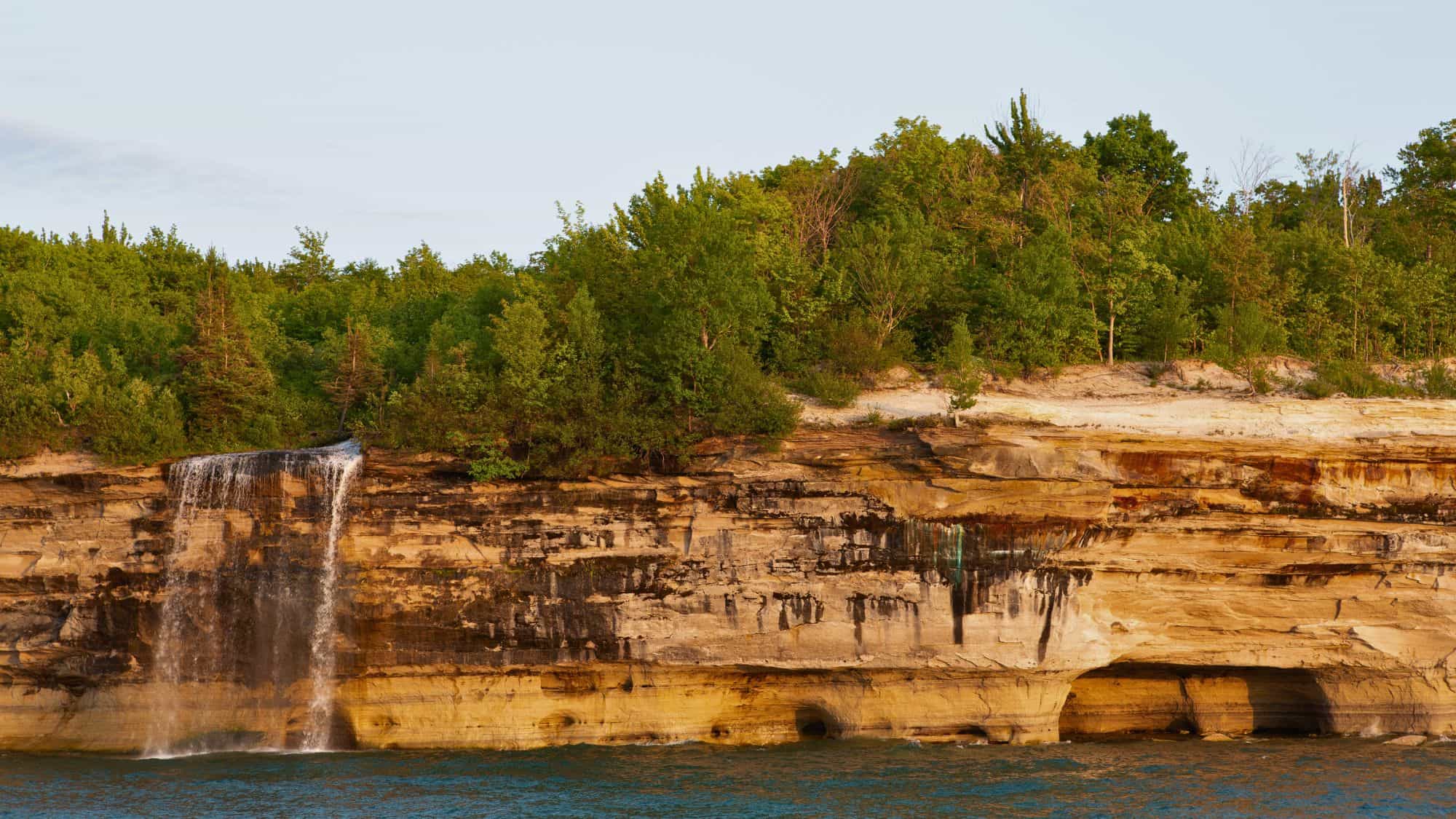 Tall, narrow waterfall plunges down a forested cliffside, surrounded by layers of golden rock and green trees on the Lake Superior shoreline.