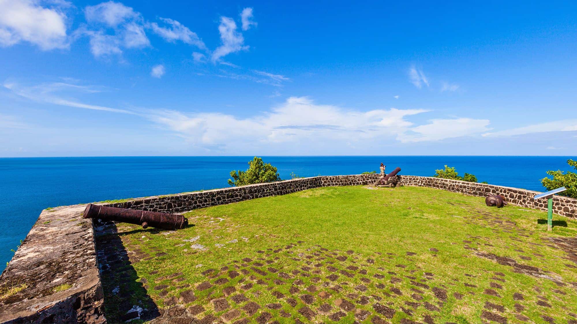 Historic stone fort walls and rusted cannons overlook the Caribbean Sea from the grassy Pigeon Island National Landmark, under a bright blue sky.