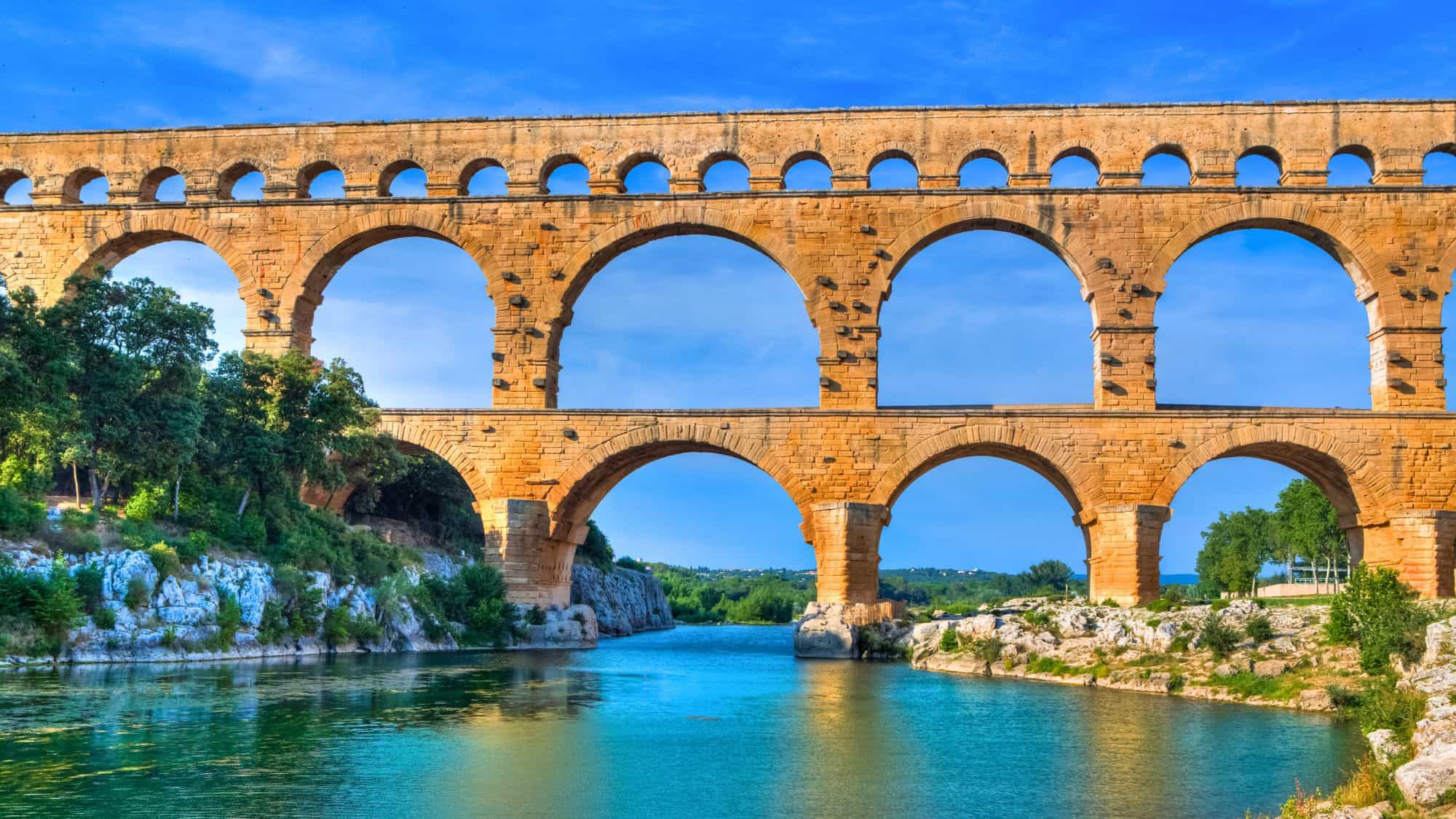 The majestic Pont du Gard aqueduct spans a tranquil blue river, with three tiers of ancient Roman arches set against a bright sky and green landscape in southern France.