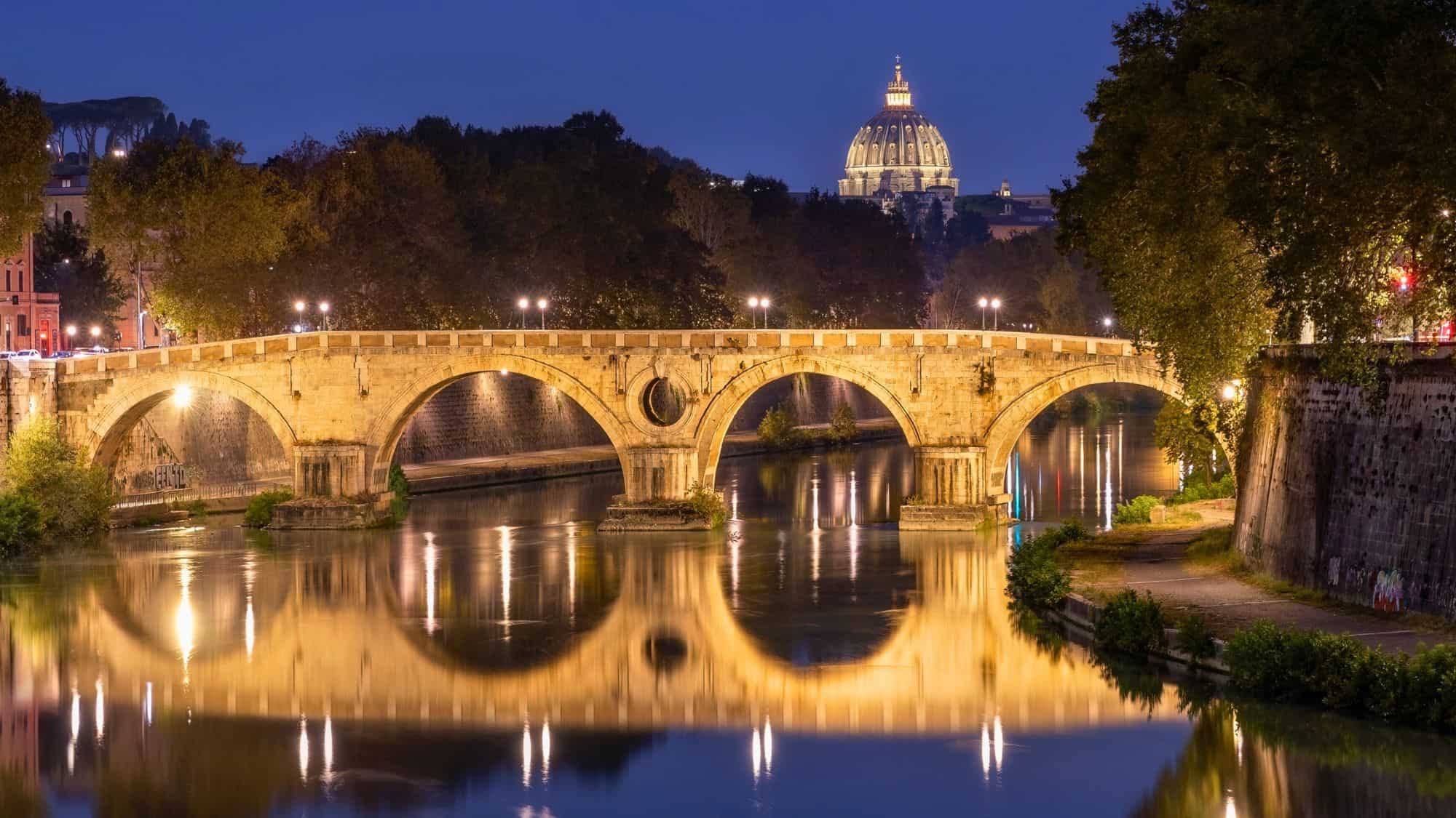 Rome, Italy - October 5, 2023 : Reflection of a historic arched bridge crossing the Tiber River with Vatican City in the background at night. Ponte Umberto I