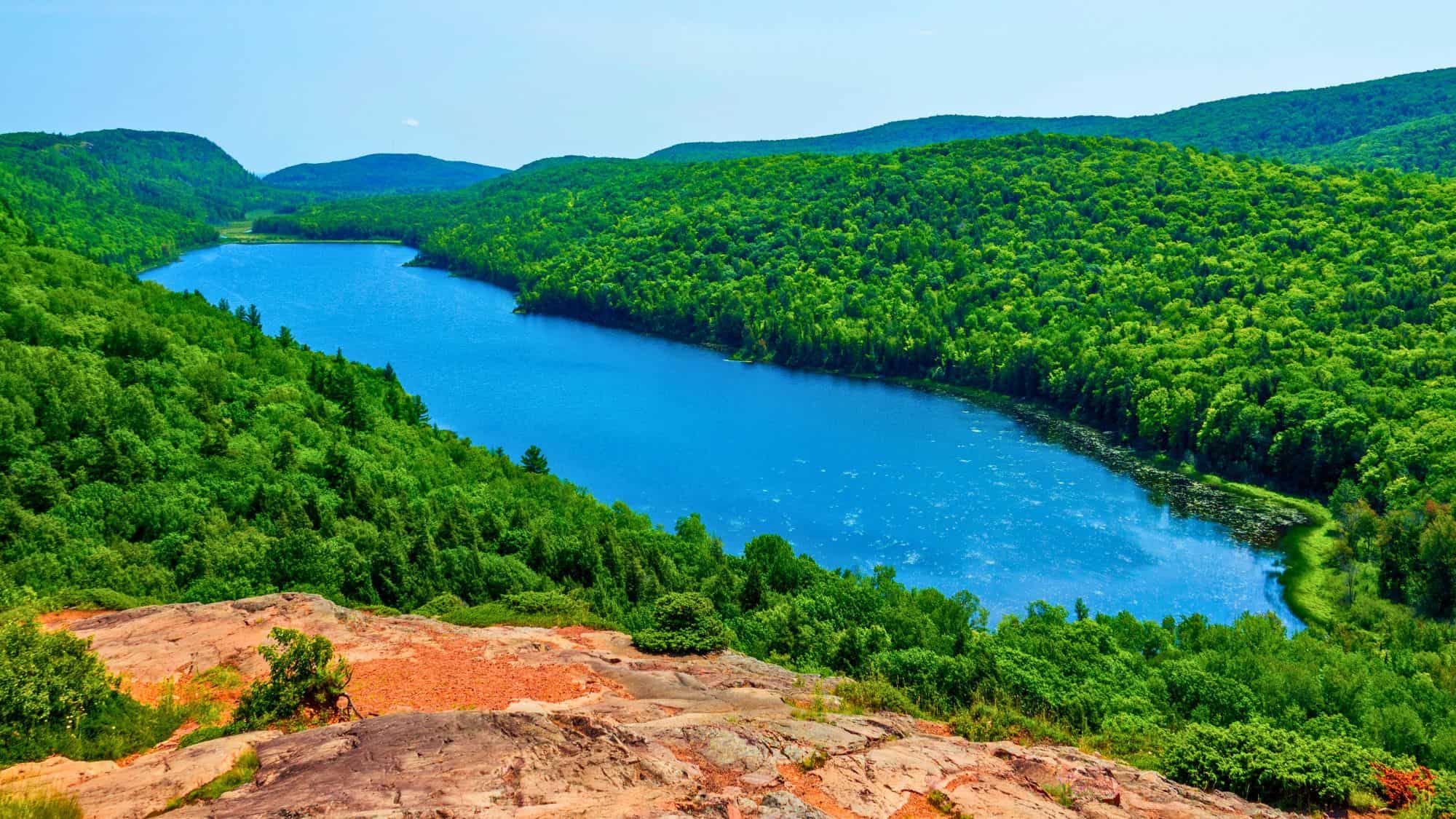 Sweeping view of Lake of the Clouds, a deep blue lake wrapped in dense, vibrant green forest and rolling hills under a clear summer sky.