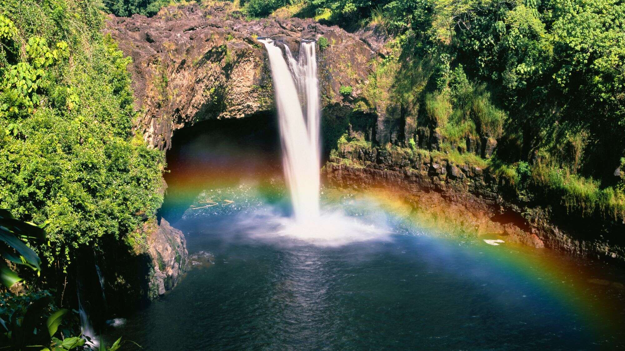 Water cascades into a pool below Rainbow Falls in Hawaii, framed by lush greenery and a vivid rainbow forming in the mist.