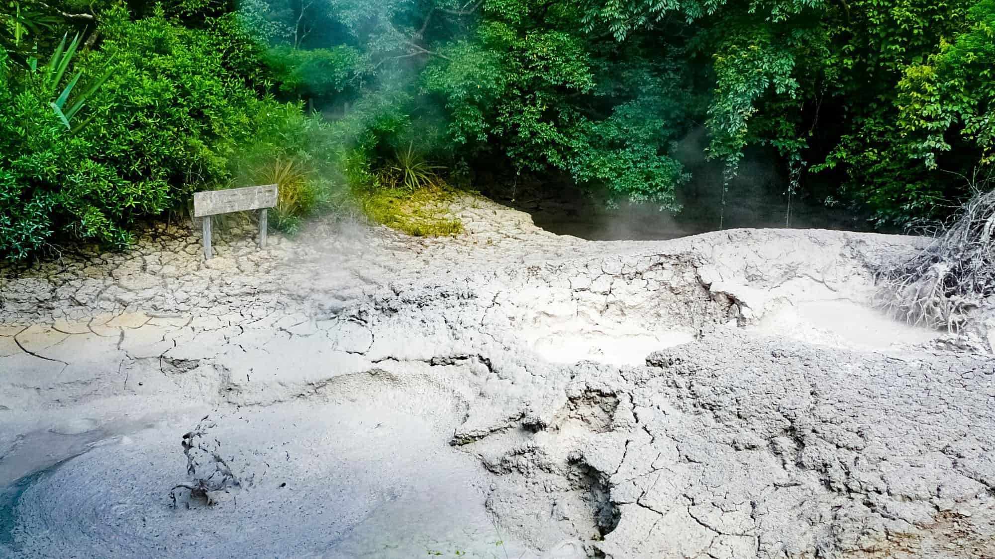A geothermal mud pot bubbles and steams within a cracked, pale gray landscape surrounded by lush, tropical greenery. The scene captures the raw power of volcanic activity in Costa Rica’s Rincón de la Vieja National Park.