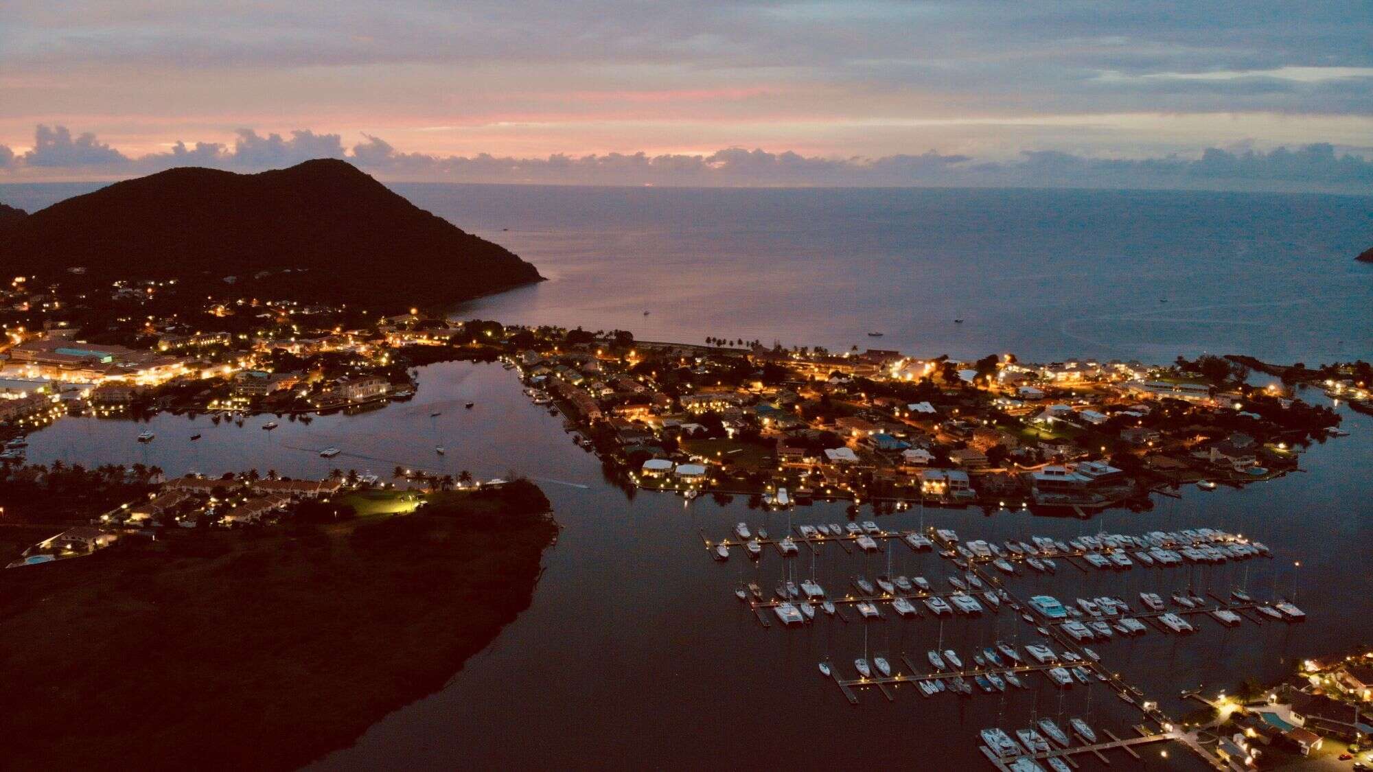 Evening aerial view of Rodney Bay lit up with golden lights, with a marina full of docked boats and a mountainous silhouette against the sea.