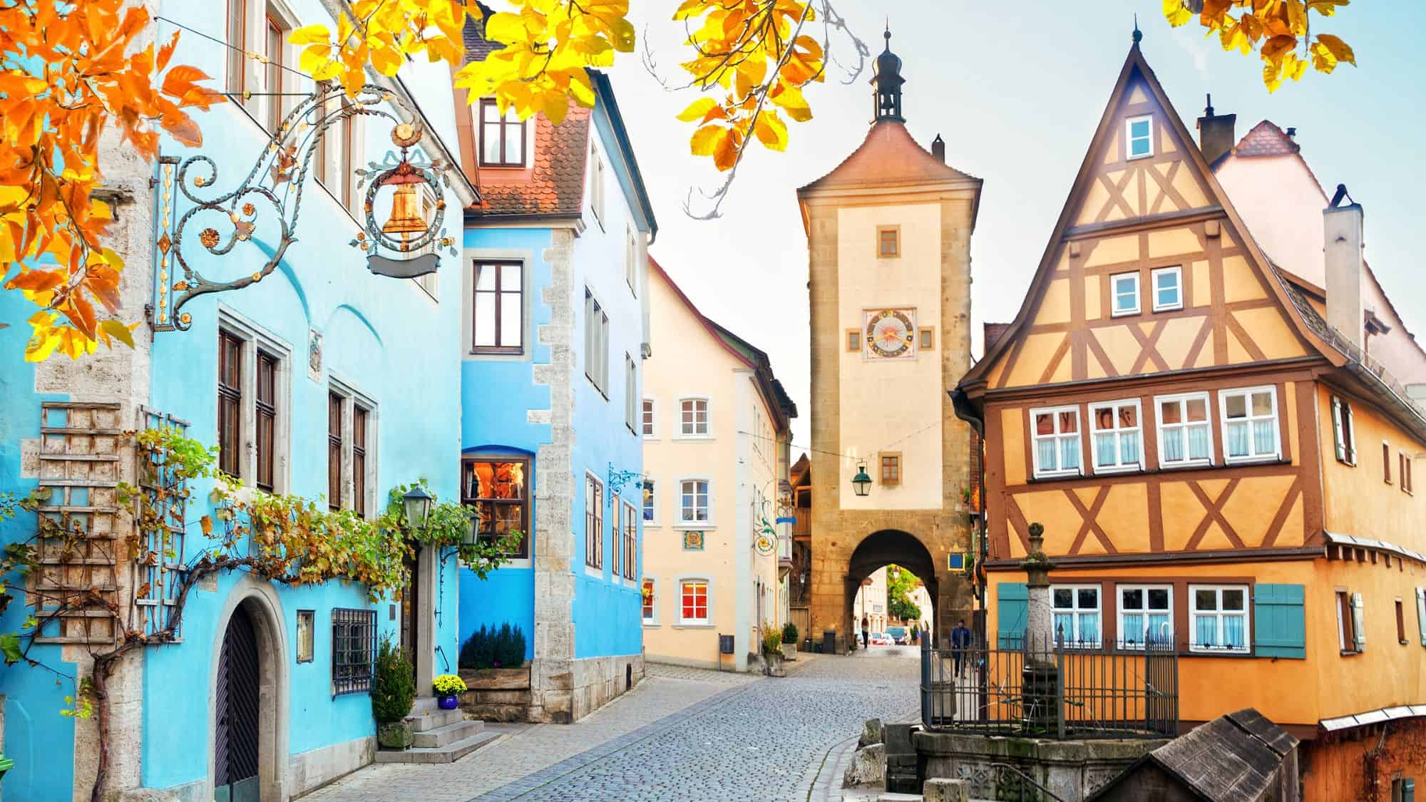 A charming medieval street lined with colorful half-timbered houses and a stone clock tower, framed by golden autumn leaves, in the fairytale town of Rothenburg ob der Tauber.