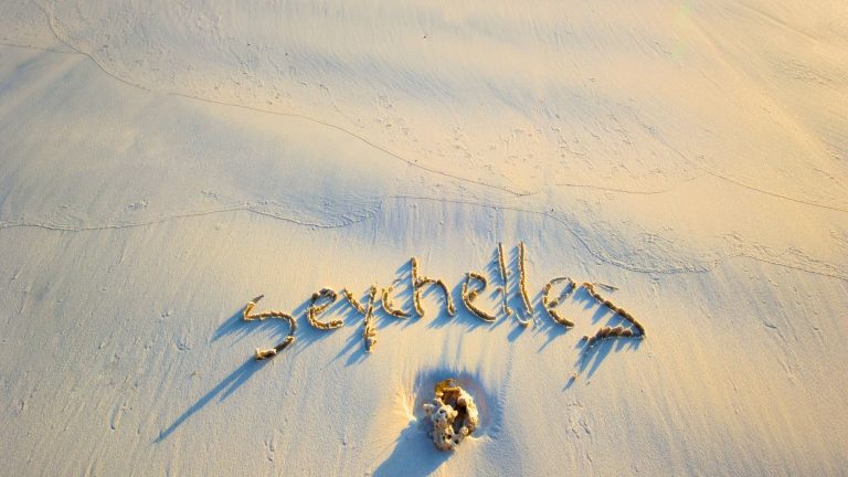 The word "Seychelles" is written in the smooth golden sand near a piece of coral, captured during the soft lighting of early morning or late afternoon.