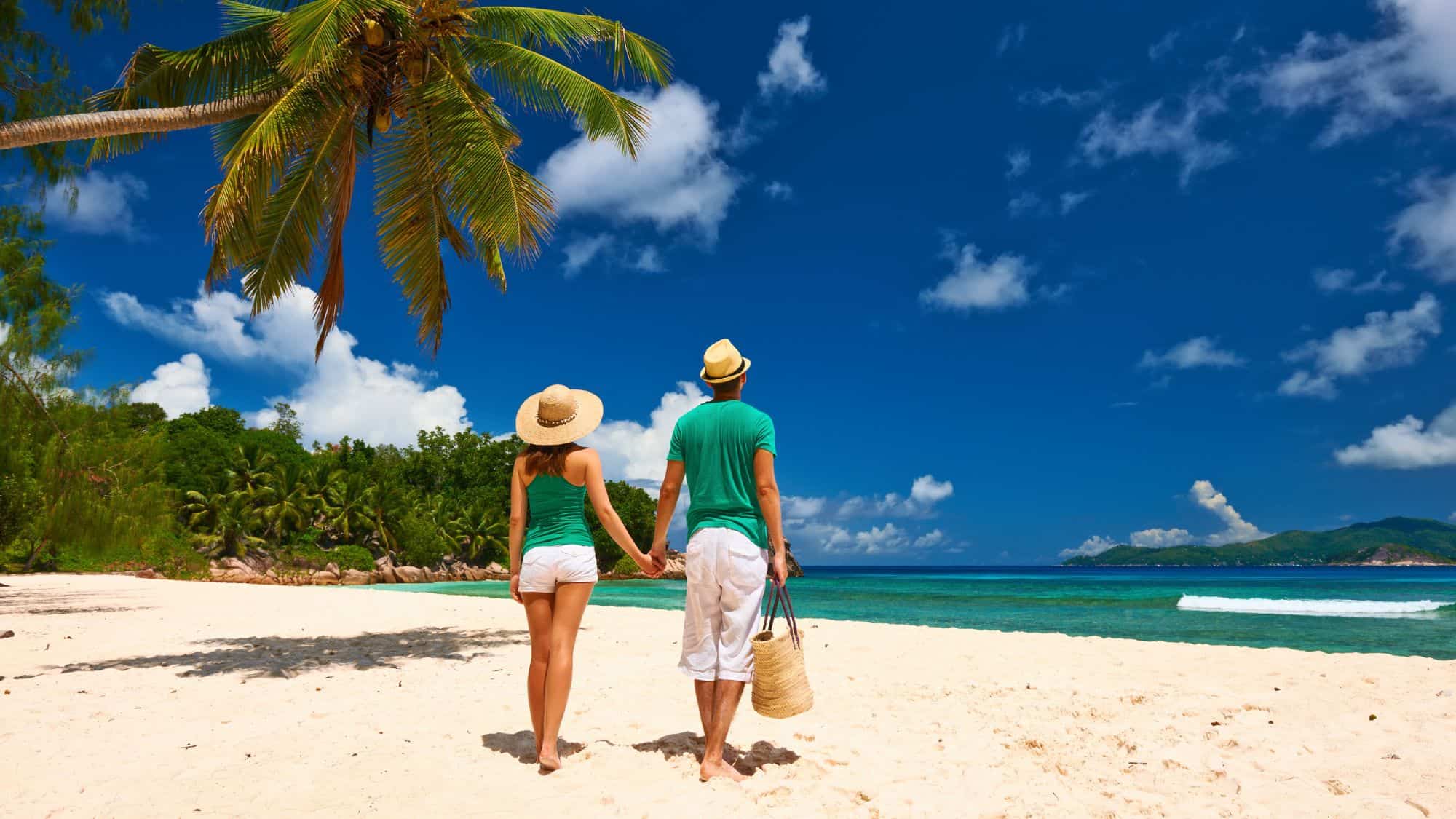 A couple wearing sunhats and casual summer clothes stands hand in hand on a pristine beach, facing the turquoise ocean under a palm tree and vibrant blue sky.