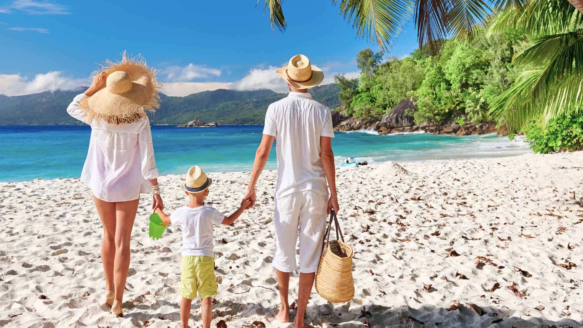 A family of three, dressed in summery white attire and straw hats, walks barefoot across the sandy shore toward the turquoise waves of a tropical beach in Seychelles, framed by lush greenery and swaying palm trees.