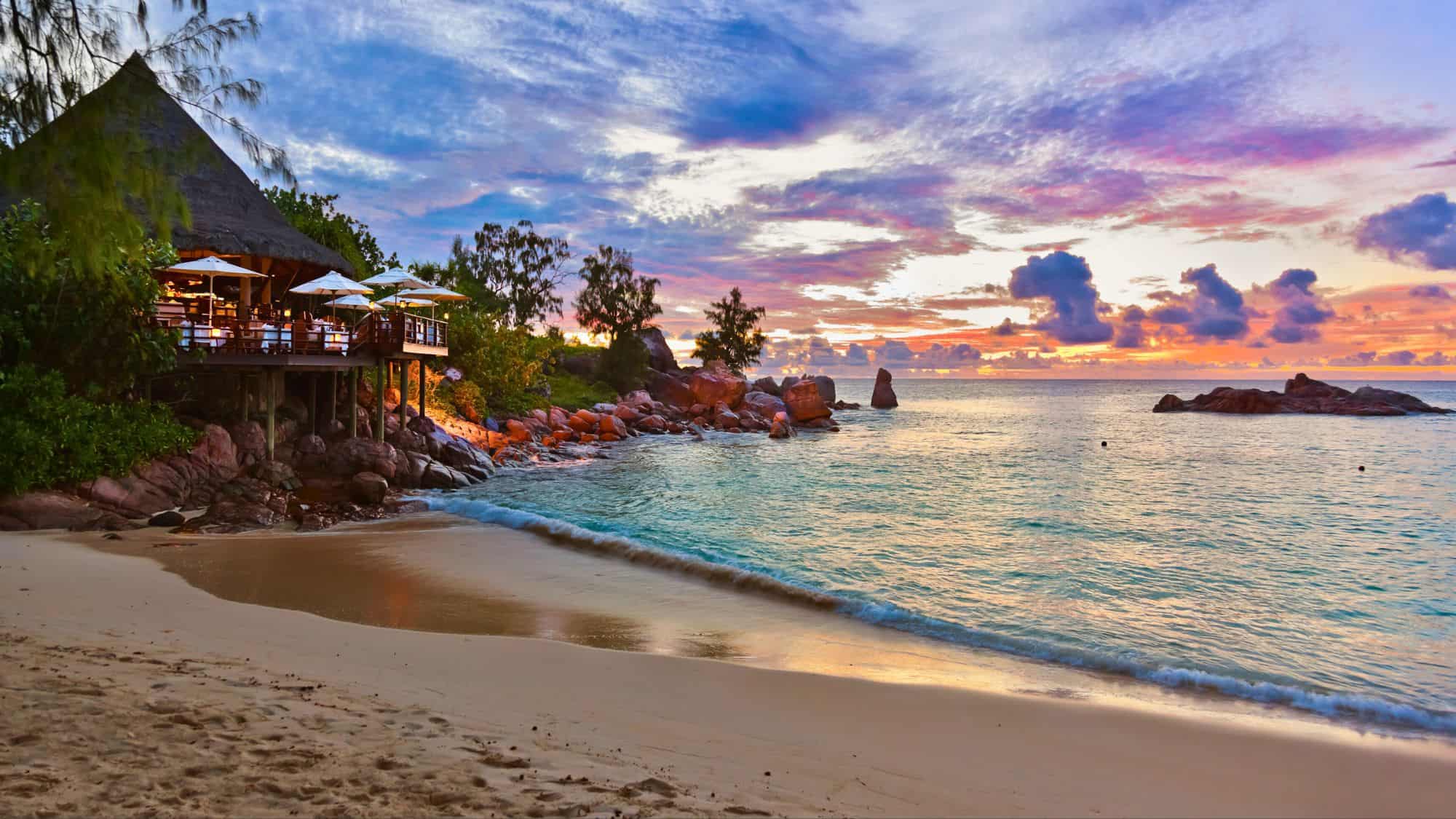 A romantic beachfront restaurant with glowing lights sits on rocky terrain beside a gently lapping ocean, under a pastel-hued sunset sky.