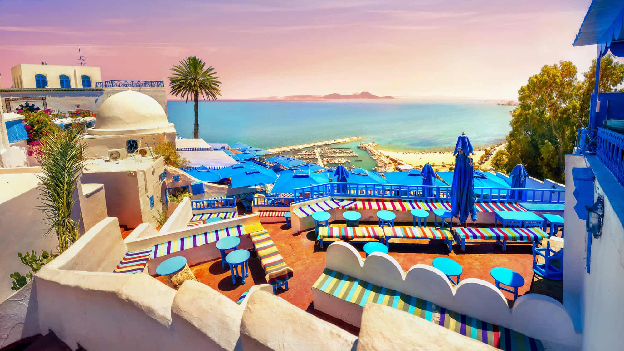 A stunning view of Sidi Bou Said’s whitewashed buildings with bright blue accents, colorful striped seating, and blue umbrellas overlooking the Mediterranean Sea at sunset.