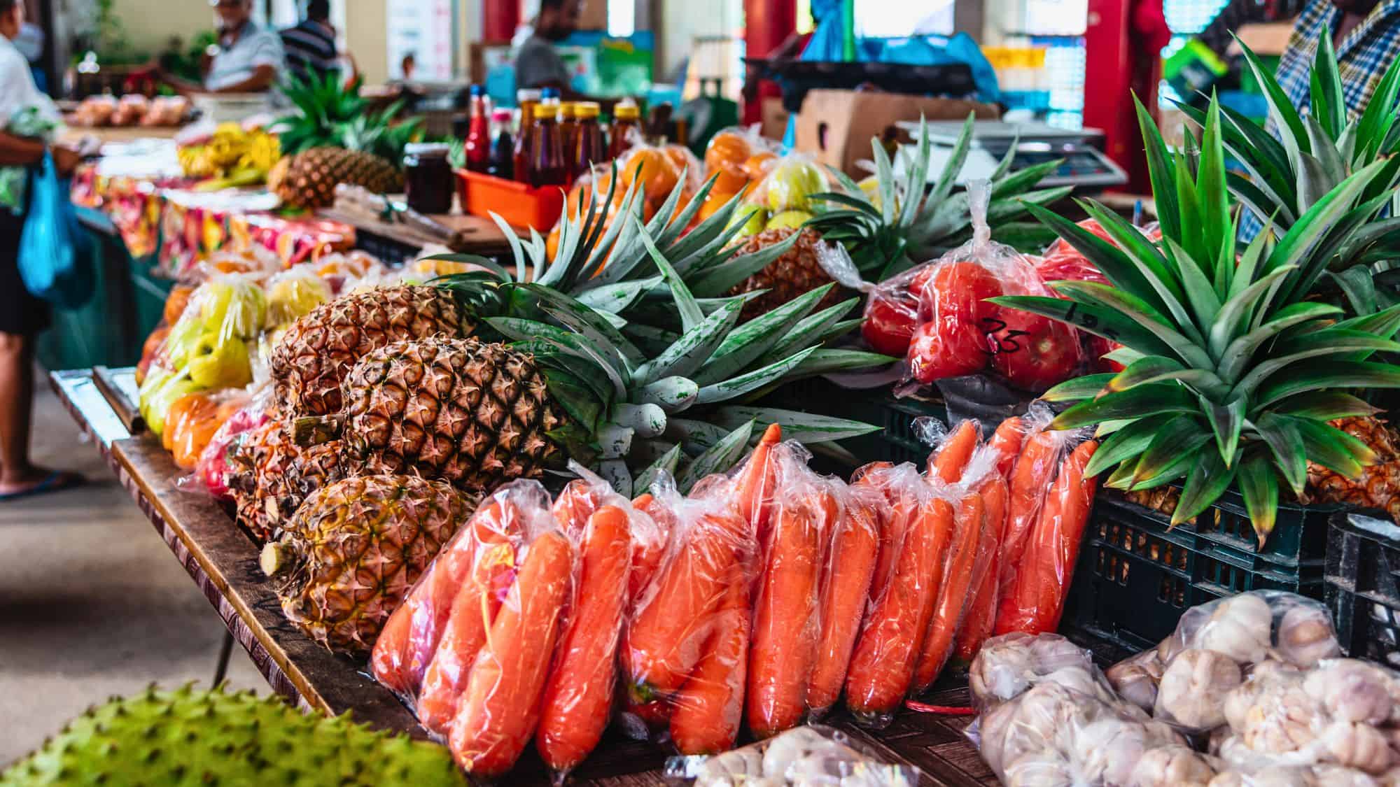 A vibrant produce stall at a Seychellois market overflows with pineapples, carrots, papayas, and other tropical fruits and vegetables, with shoppers browsing in the background.