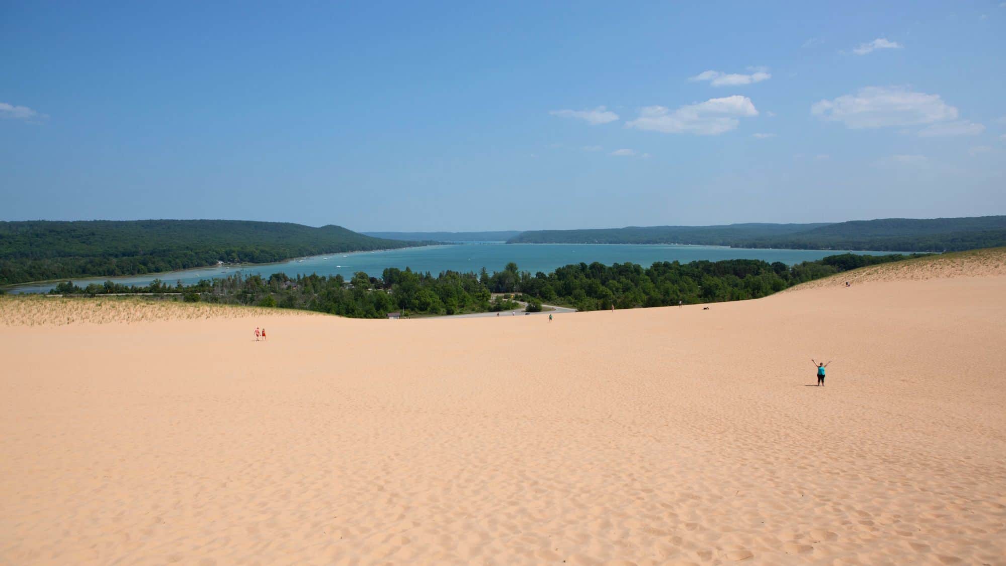Expansive sand dune stretching out toward a forested area and blue lake in the distance, with tiny people dotting the landscape for scale.