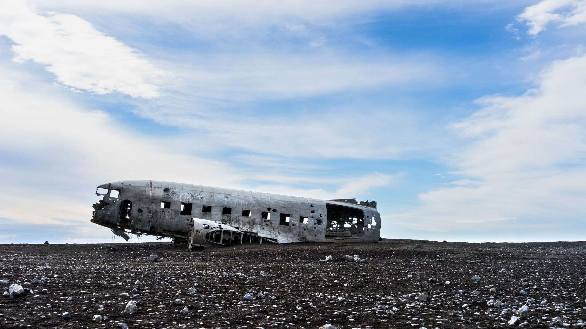 A weathered, abandoned DC-3 airplane rests on the black sand beach of Sólheimasandur in Iceland, surrounded by scattered stones under a partly cloudy sky.