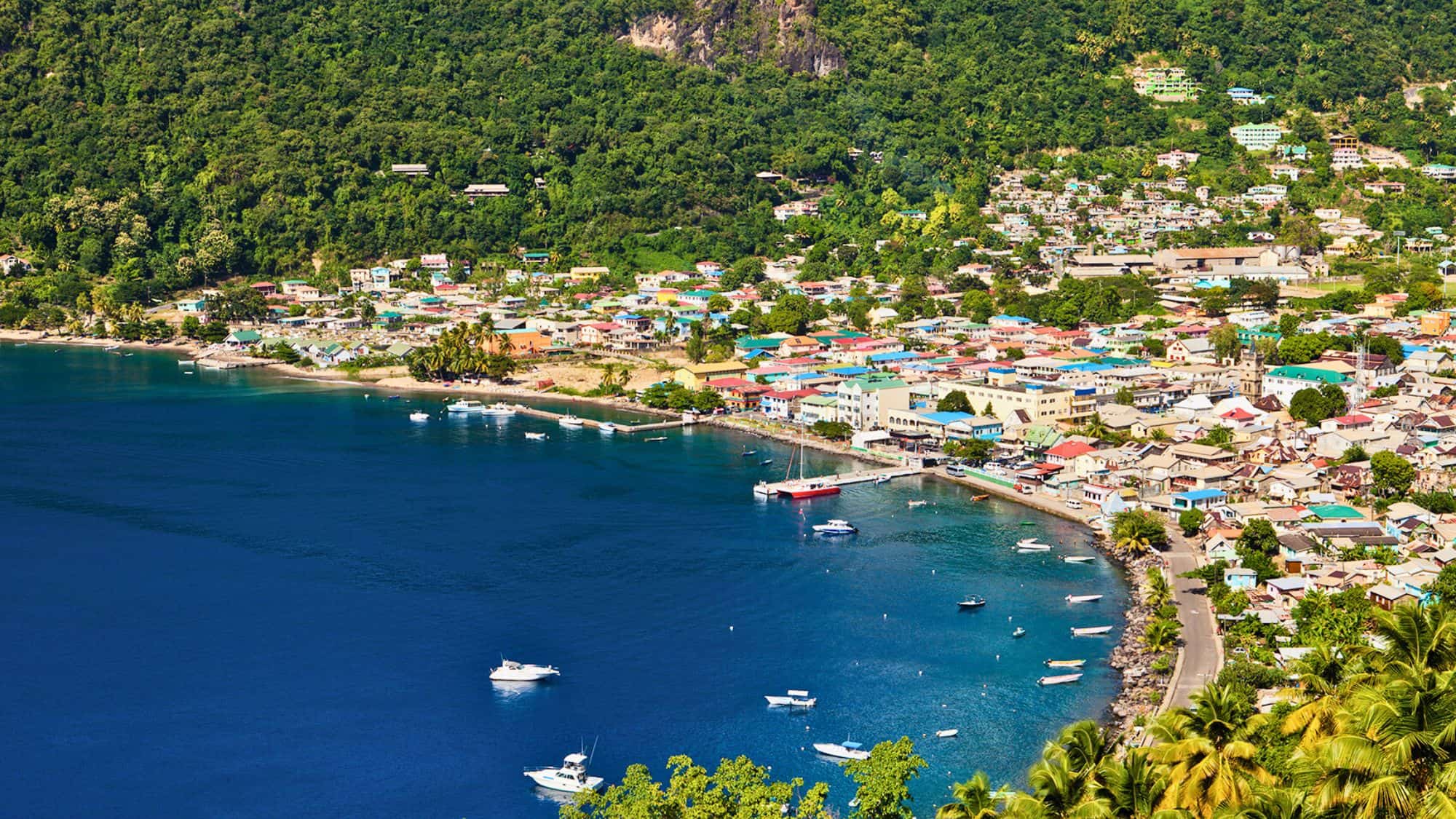 Colorful coastal town of Soufrière nestled between rainforest-covered hills and a curved shoreline, dotted with small boats in calm blue waters.
