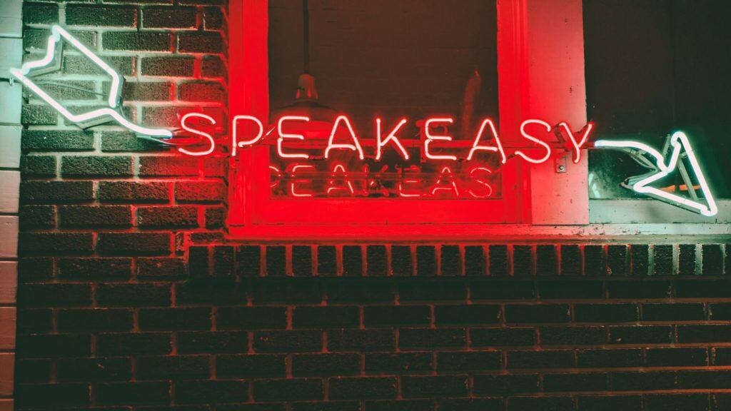 A red neon sign reading “SPEAKEASY” glows in a window, bordered by white neon arrows pointing inward, mounted on a dark brick wall and casting a retro vibe.