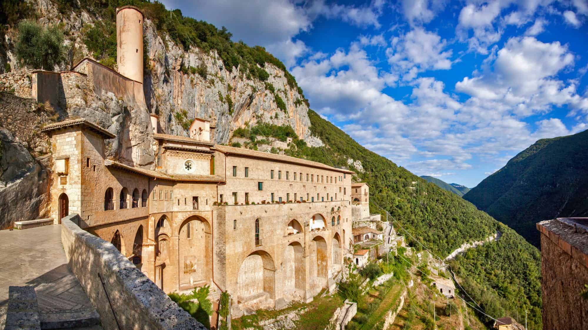 A historic monastery built into the mountainside at Subiaco, Italy, with arched foundations clinging to rocky cliffs and green hills all around.