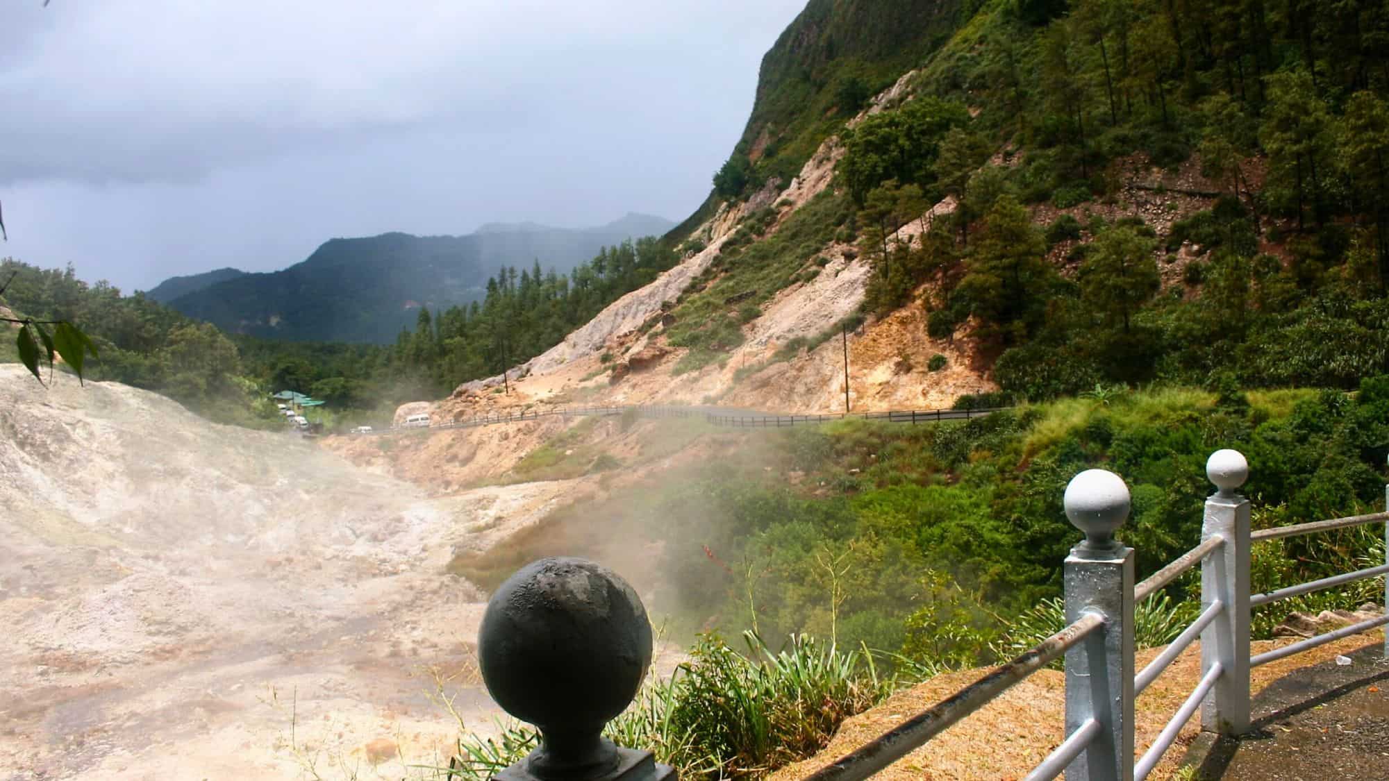 Geothermal landscape at Sulphur Springs Park, featuring steaming rocky terrain, mineral-stained slopes, and a winding road along a forested hillside.