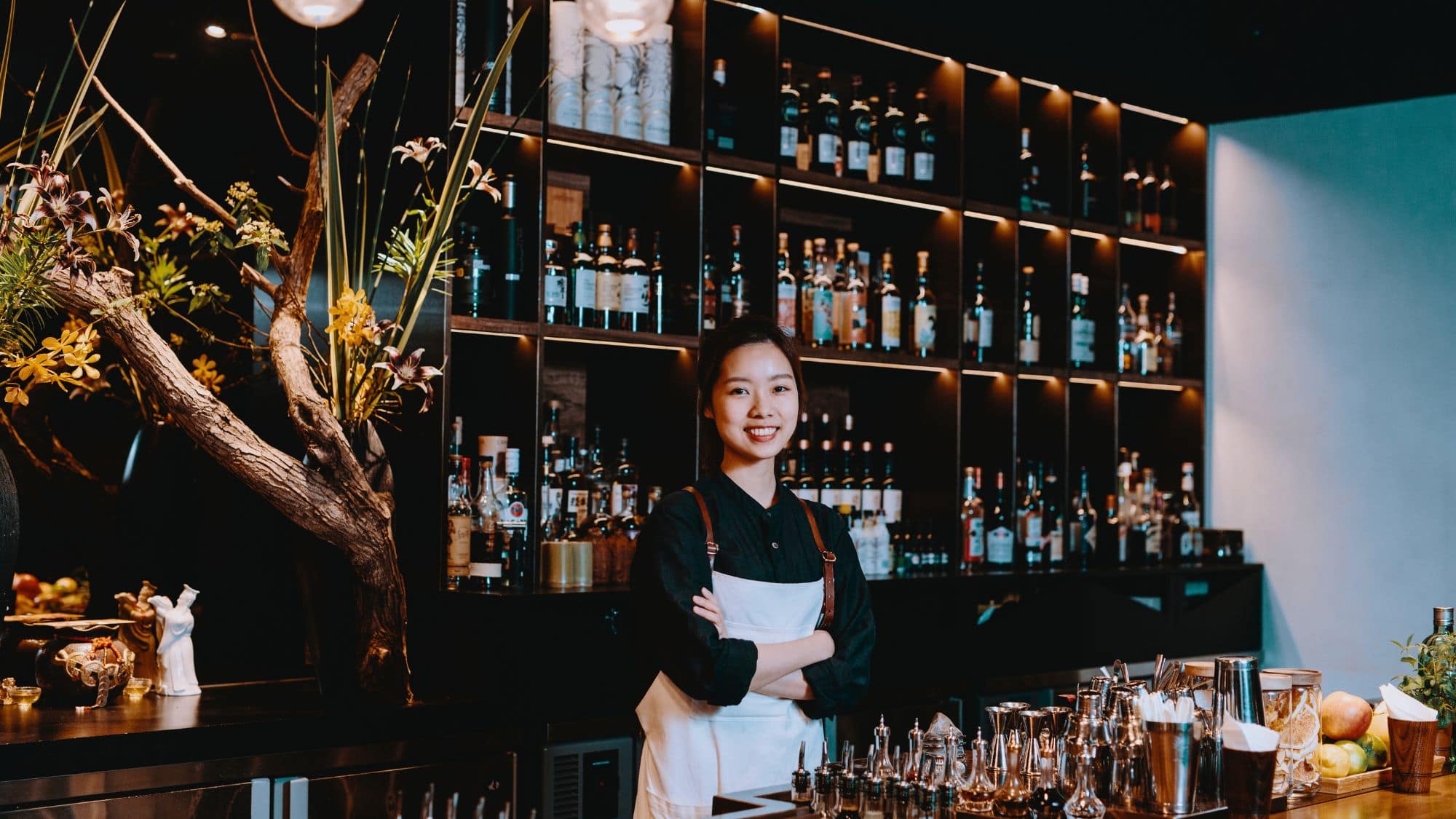 A bartender stands confidently with folded arms behind a sleek bar in Taiwan, surrounded by elegant bottles, fresh garnishes, and modern floral decor.