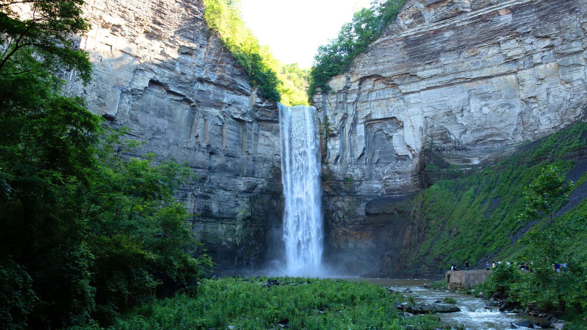 A dramatic vertical waterfall plunges down a sheer cliff face into a green basin below, framed by lush greenery and layered rock walls.