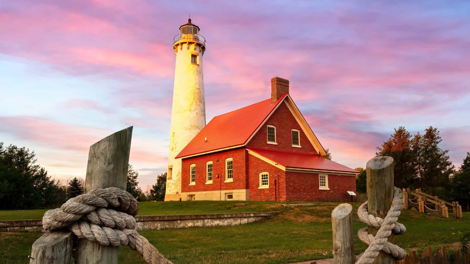 Classic white lighthouse with a red roof and tall tower stands against a pastel sky, surrounded by grassy lawn, wooden posts, and nautical ropes.