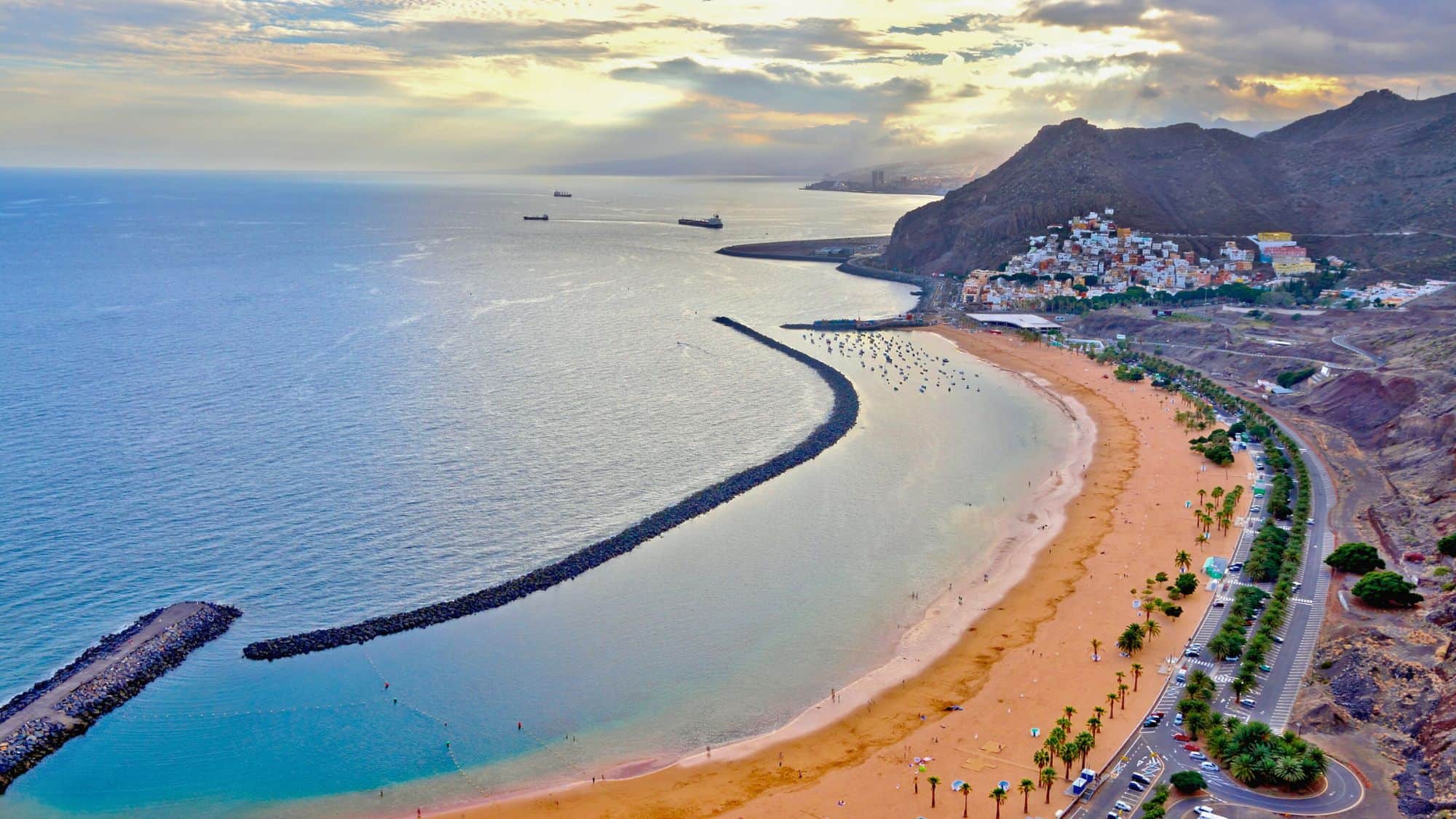 An expansive aerial view of Las Teresitas Beach in Tenerife, with golden sand, calm turquoise water protected by breakwaters, palm trees lining the promenade, and a hillside village tucked at the base of surrounding cliffs.