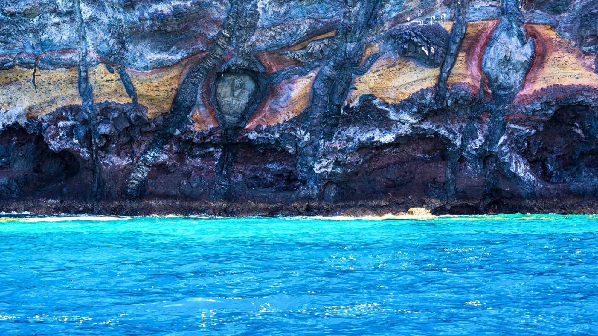 A close-up of the colorful stratified volcanic rock of a Tenerife sea cliff, showcasing vivid layers of red, orange, and black rock above the vibrant blue ocean.