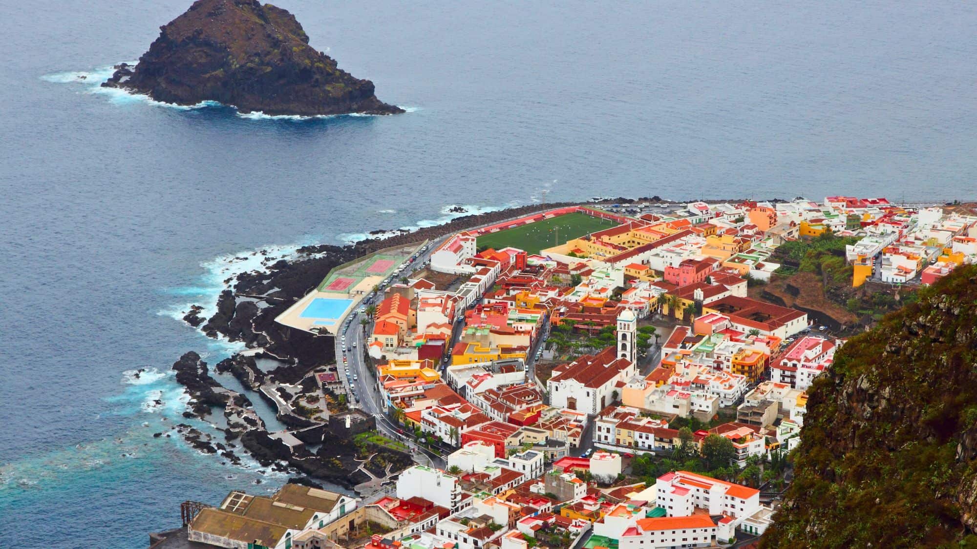 A panoramic aerial view of a bustling coastal city in Tenerife with high-rise buildings, palm-lined streets, and a large lagoon-style pool by the ocean, with Mount Teide and surrounding hills in the hazy distance.
