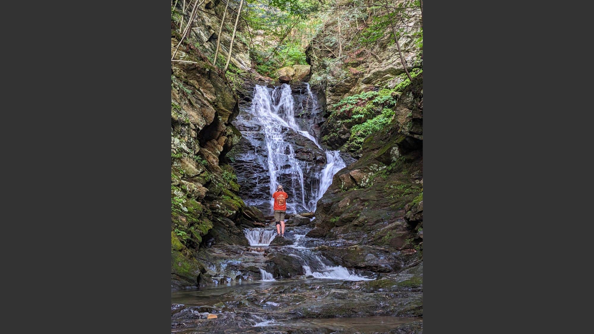 A man in an orange shirt stands at the base of a narrow, multi-tiered waterfall nestled between steep rock walls, framed by trees and mossy boulders.