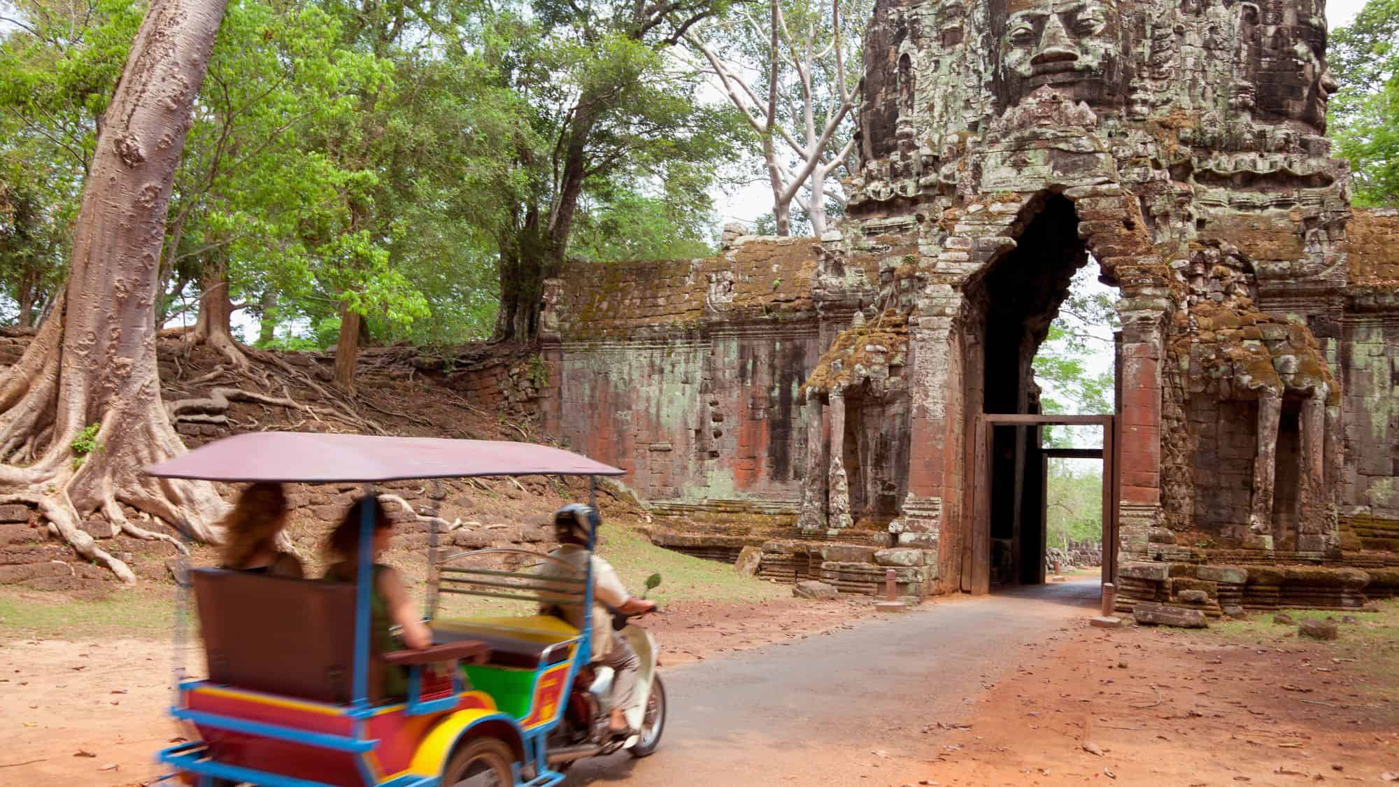 A colorful tuk-tuk carrying tourists approaches the ancient gate of Angkor Thom in Cambodia, surrounded by giant tree roots and moss-covered stone architecture.