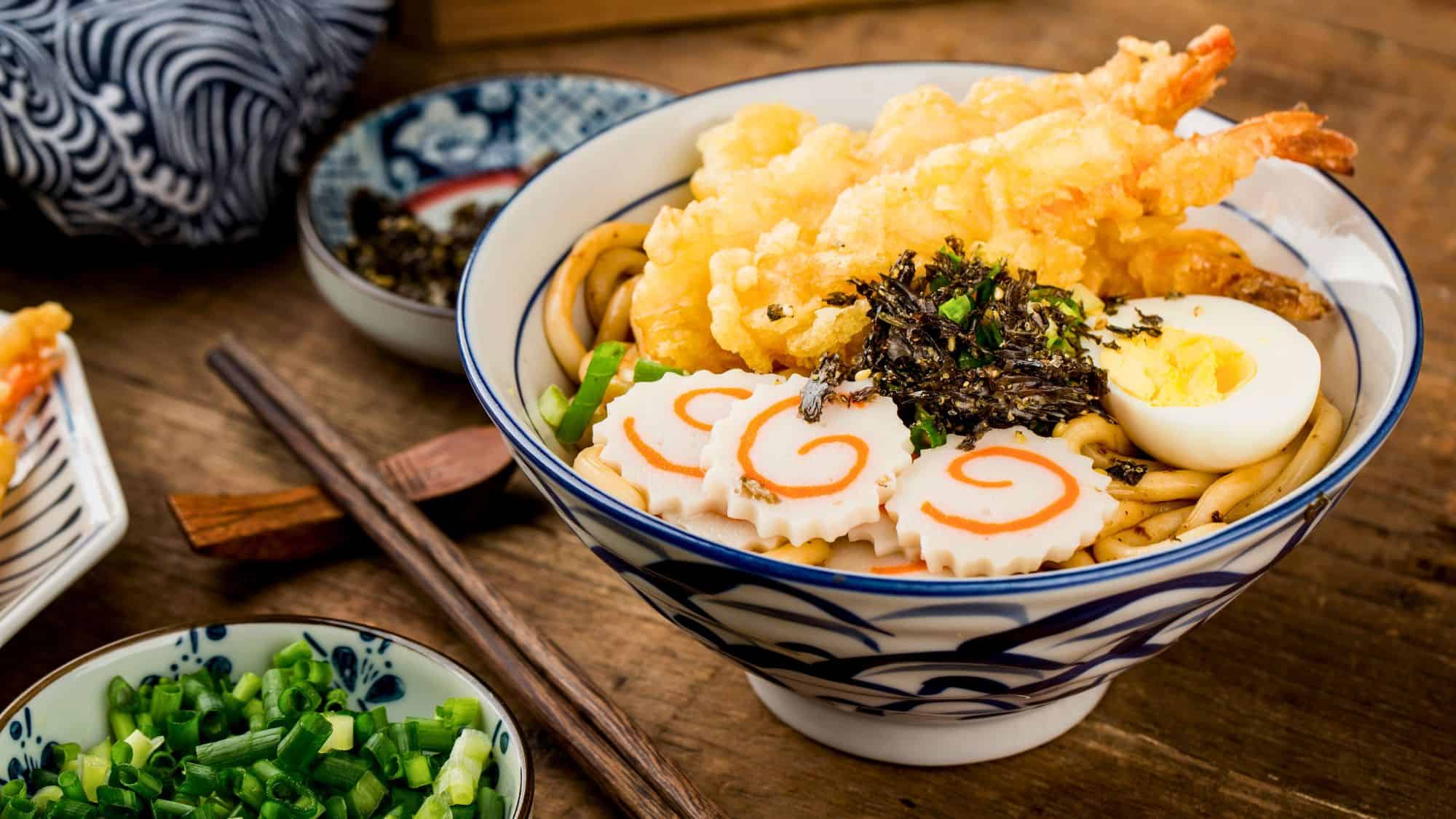 A vibrant bowl of udon noodles topped with shrimp tempura, sliced fish cake with spiral designs, half a boiled egg, seaweed, and chopped green onions, arranged on a wooden table with side dishes.