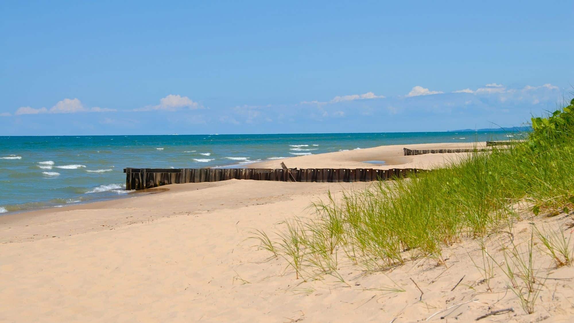 Peaceful stretch of golden sand along Lake Michigan, with rolling dunes, green grasses, and gentle waves washing up under a blue sky.