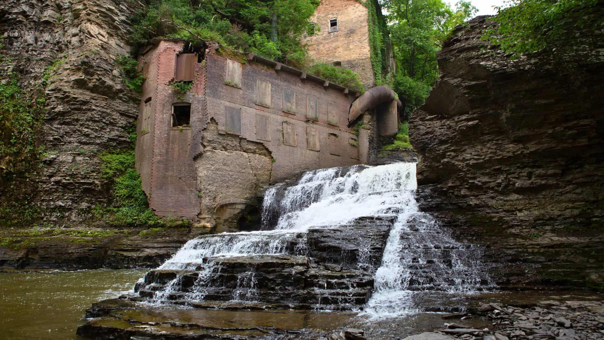 A waterfall spills over stone steps next to an abandoned brick mill with boarded-up windows, surrounded by steep cliffs and overgrown vegetation.