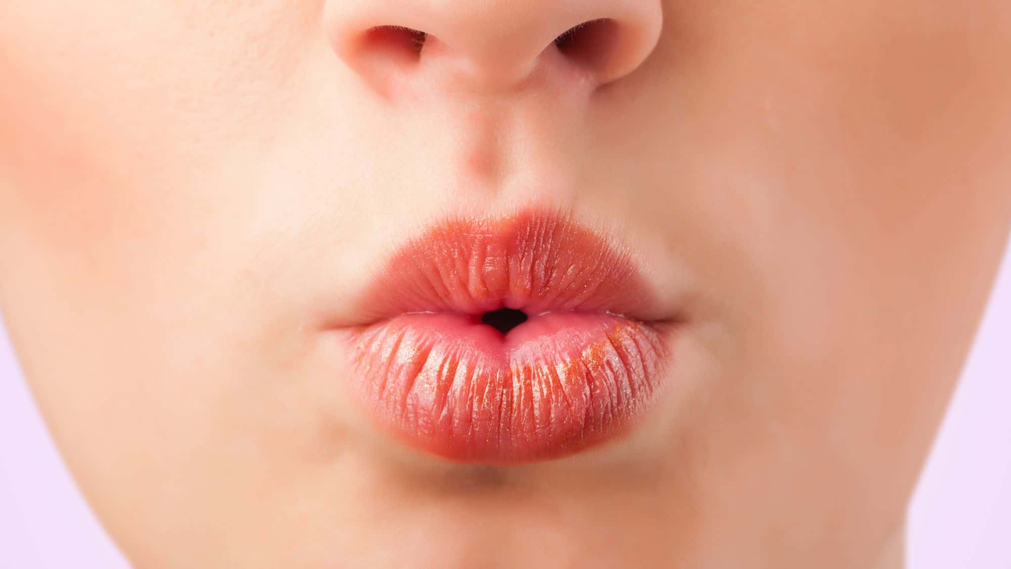 Extreme close-up of puckered, lightly glossed lips whistling, showing skin texture and soft lighting on a pale pink background.