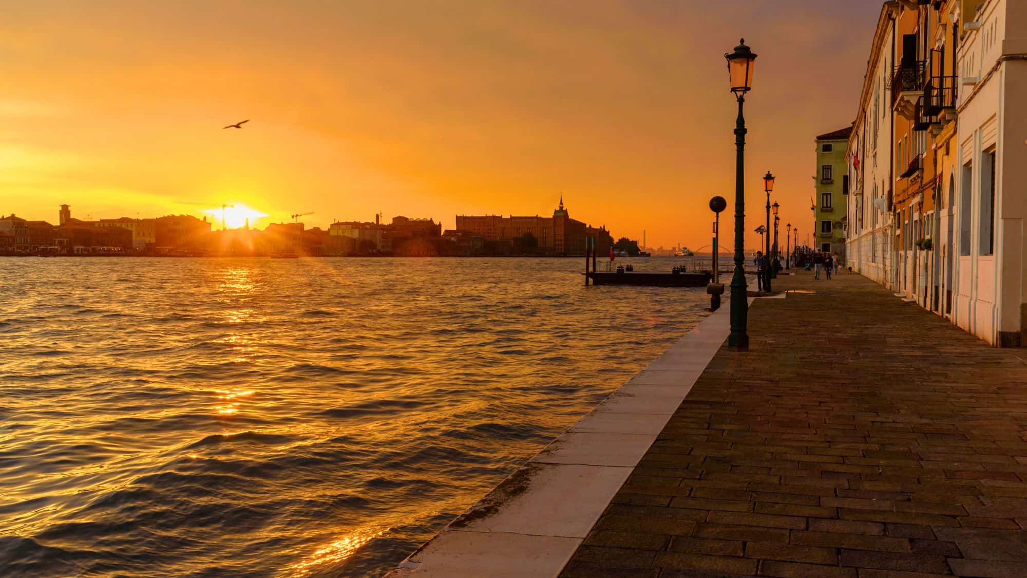 A serene sunset along the Zattere promenade, with golden light reflecting on the water and silhouetted lampposts lining the waterfront walkway.