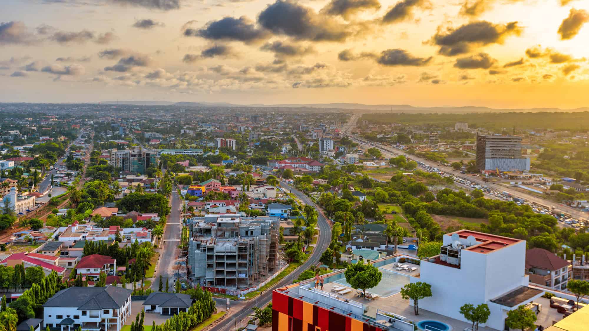 Panoramic view of Accra, Ghana at sunset, showing a sprawling cityscape with a winding road, construction sites, and modern buildings beneath golden-orange clouds.