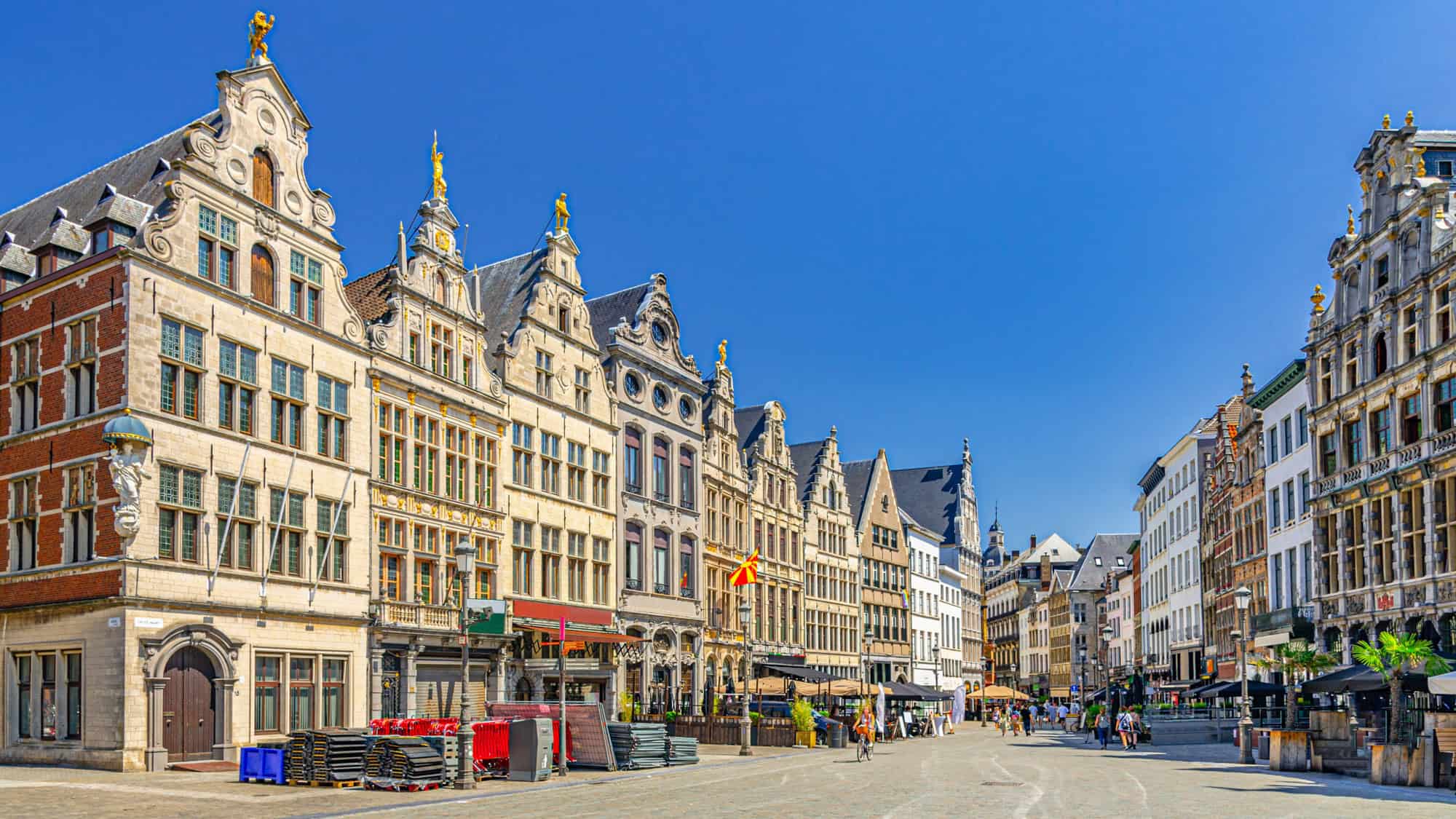A wide cobblestone square in Antwerp, Belgium, is bordered by rows of ornate gabled buildings with gilded statues and a deep blue sky overhead, showcasing classic Flemish architecture.