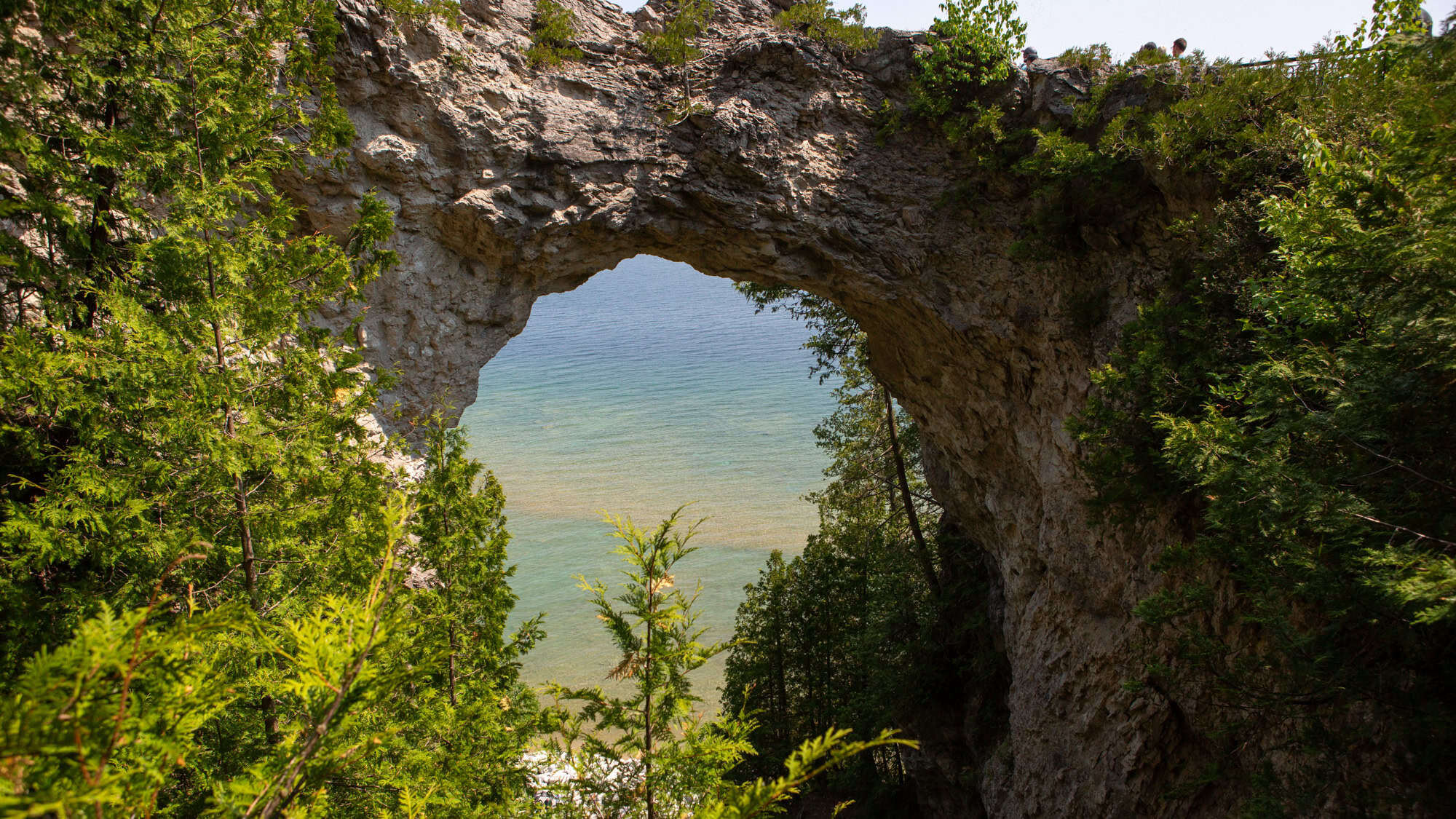 Natural limestone arch known as Arch Rock on Mackinac Island, framed by lush green trees with Lake Huron’s blue and turquoise waters visible through the opening.