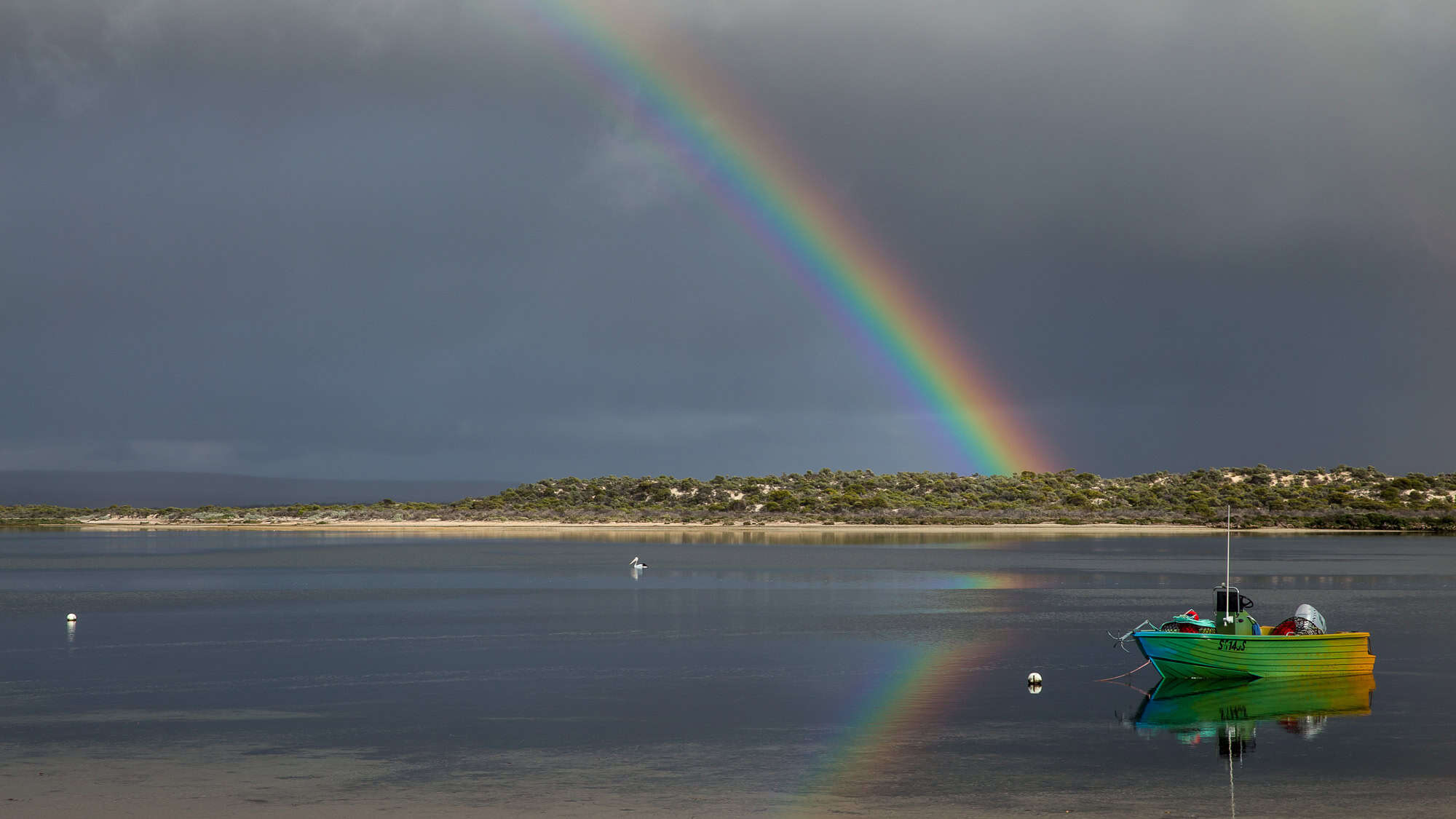 A tranquil bay scene featuring a vibrant green and yellow fishing boat floating near the shore, with a bright rainbow arching over a cloudy sky.