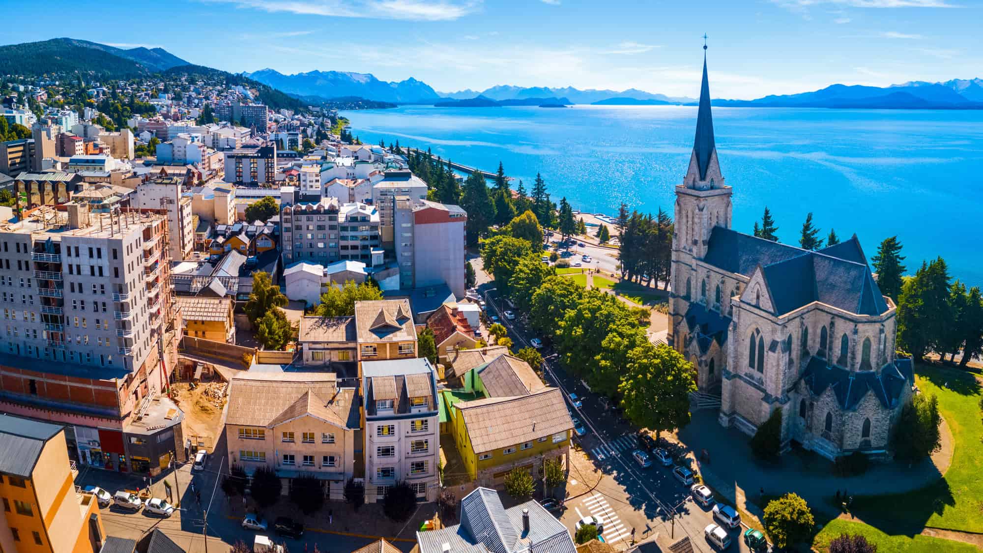 A panoramic view of San Carlos de Bariloche shows a striking lakeside cityscape with a mix of modern and alpine-style buildings. A large stone cathedral with a tall spire stands prominently near the shimmering blue lake, backed by the Andes mountains under a clear sky.